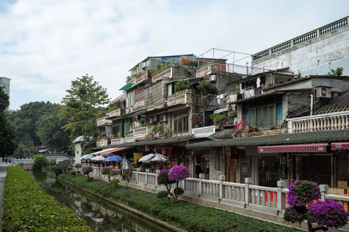 Traditional houses with balconies and lush plants line a canal in the historic area of Panyu, with a pedestrian walkway and blooming flowers in the foreground.