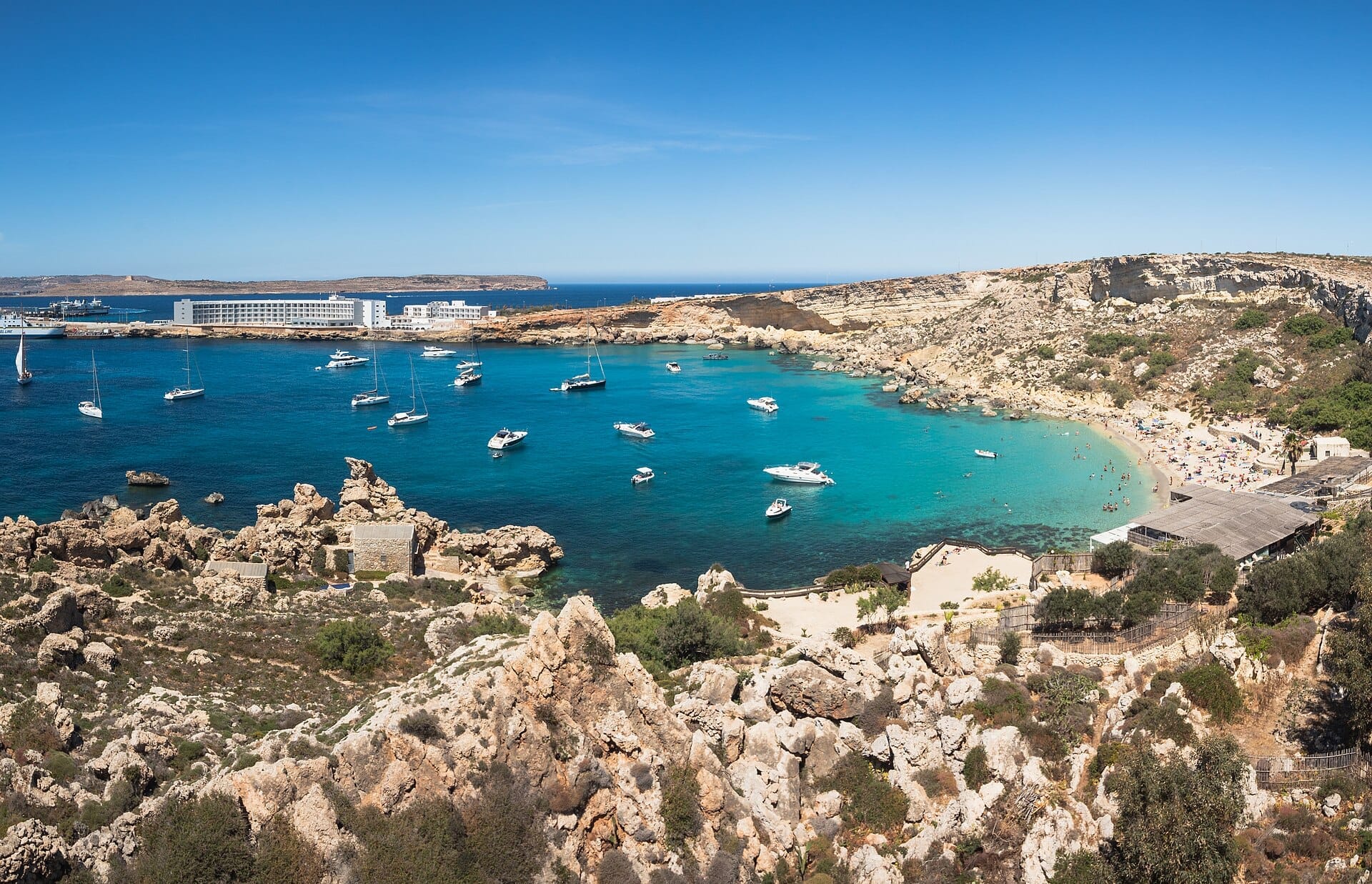 Vista panoramica di Paradise Bay, Malta, con acque turchesi, barche ancorate, costa rocciosa, spiaggia sabbiosa ed edifici vicini sotto un cielo sereno.