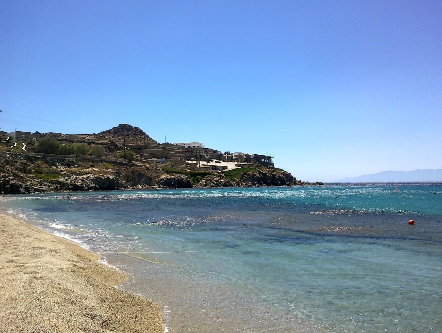 Agua azul cristalina y arena dorada bordean Paradise Beach, Mykonos, con colinas rocosas y edificios blancos visibles bajo un cielo soleado.