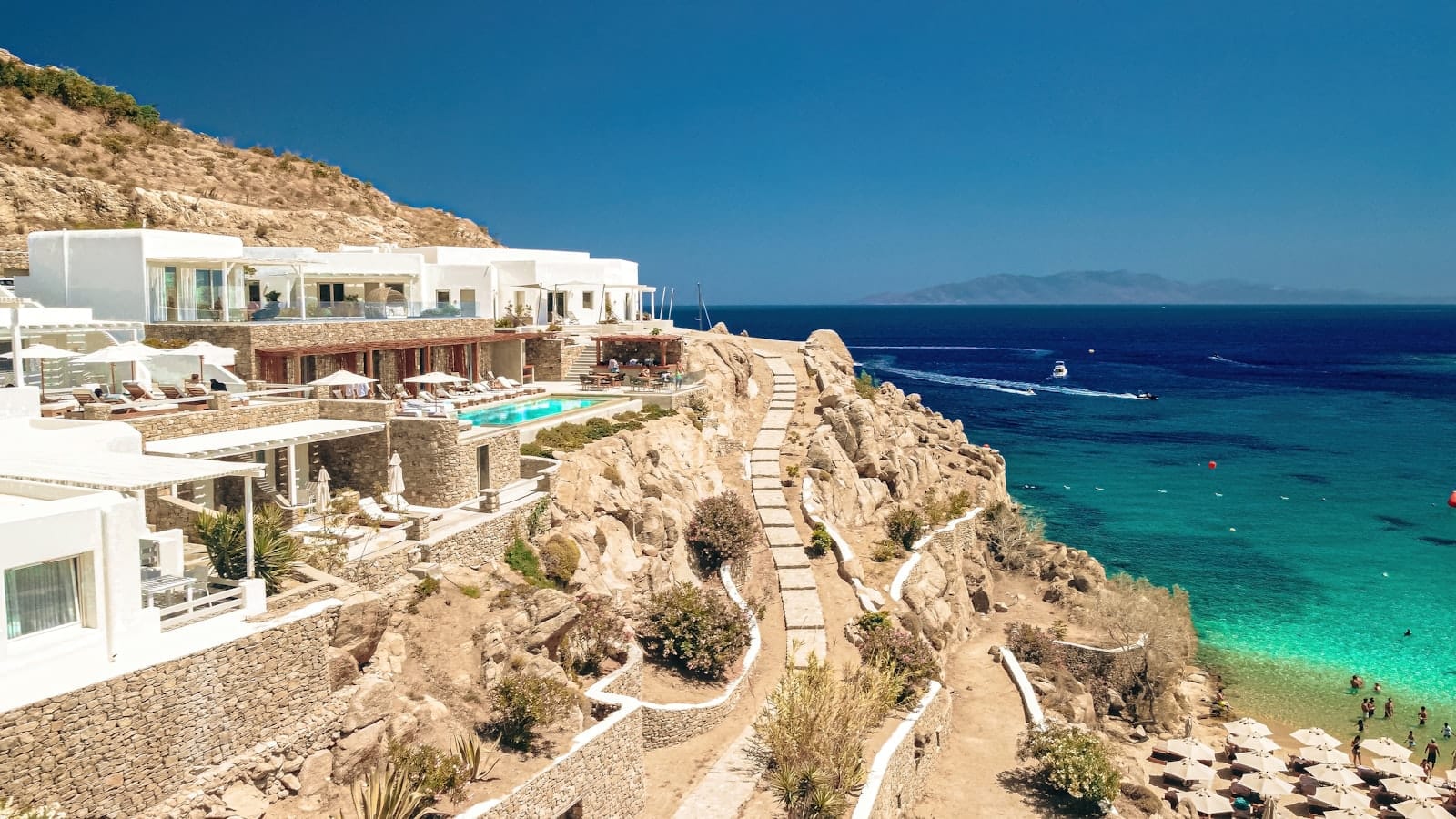 Vista desde el acantilado de Paradise o Super Paradise Beach en Mykonos con agua turquesa, tumbonas, edificios blancos de lujo y un cielo azul despejado.