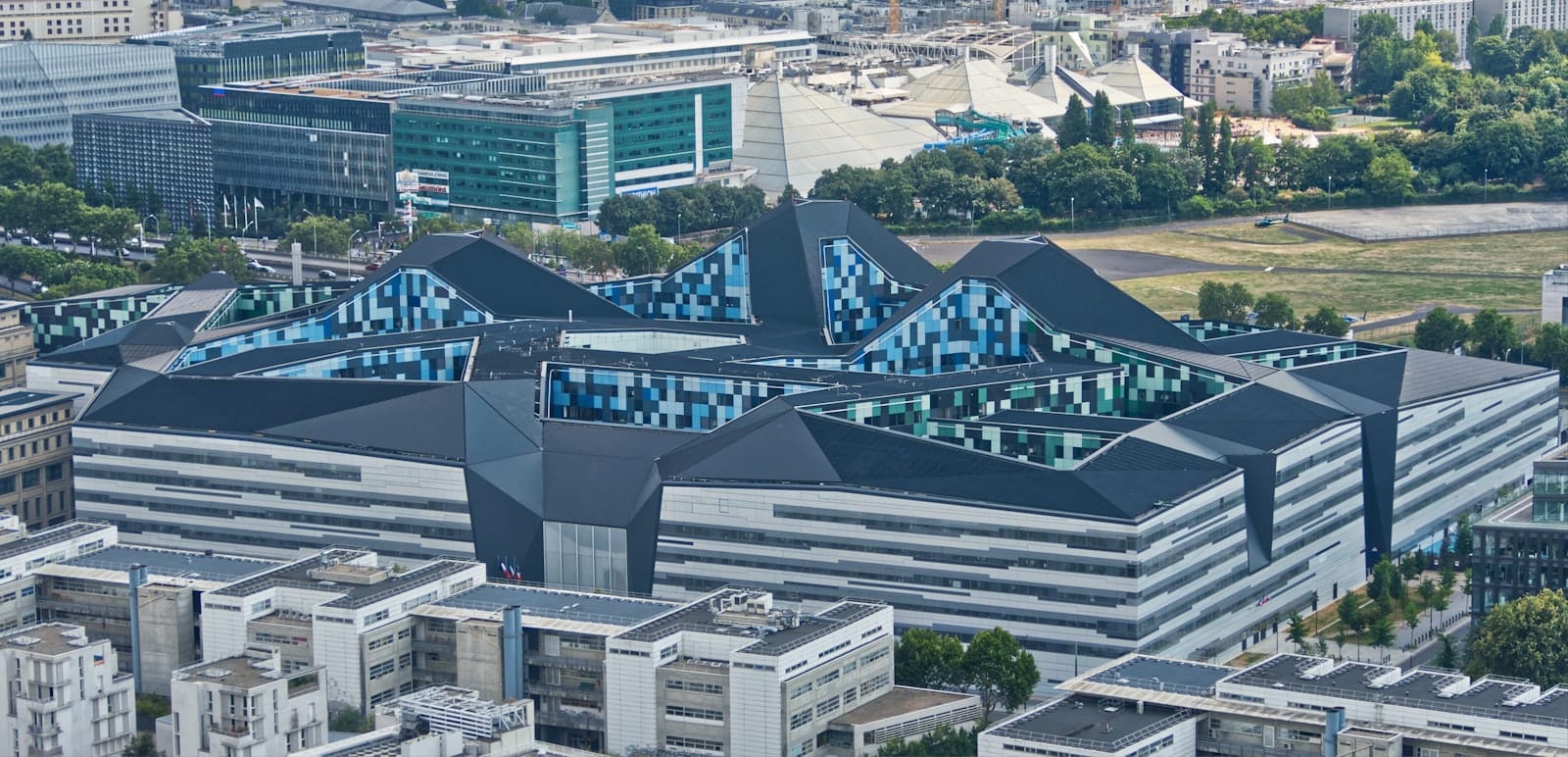 Vista aerea di un parco urbano moderno e geometrico con elementi architettonici di forte impatto, circondato dagli edifici e dagli spazi verdi di Parigi in una giornata limpida.