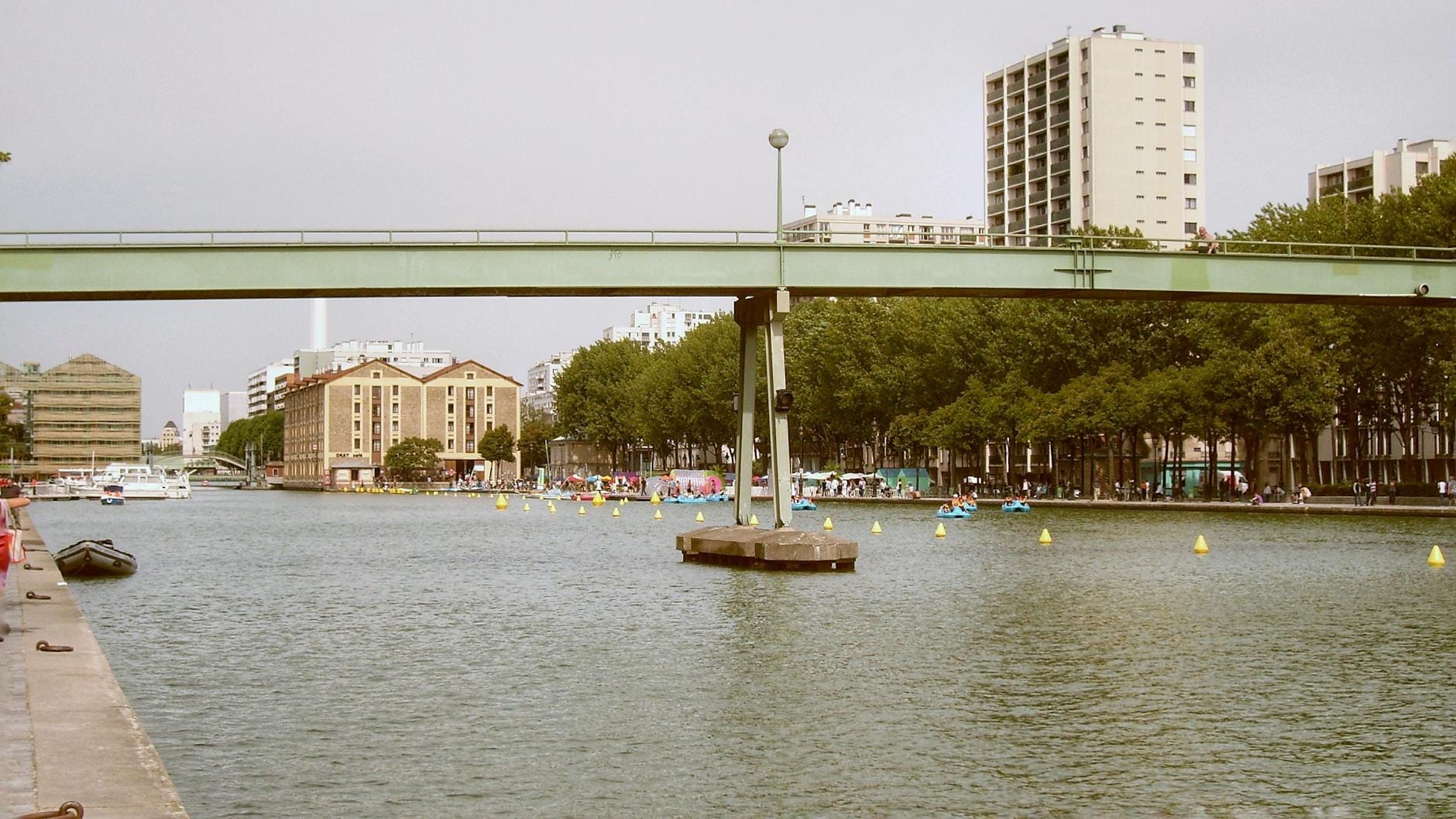 Un ampio canale in un parco urbano con un ponte pedonale, viali alberati, edifici moderni e persone che godono del parco su entrambe le sponde dell'acqua.