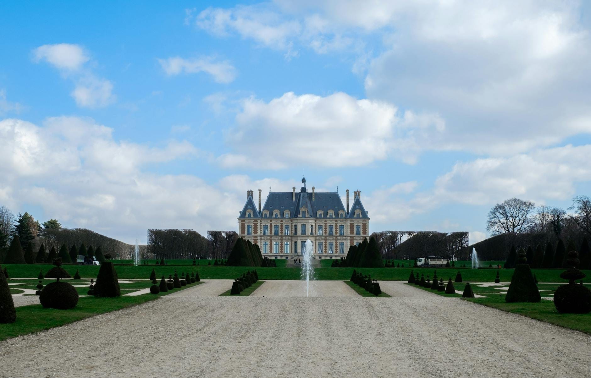 Vista ampia dello château del Parc de Sceaux incorniciato dai giardini formali alla francese, con vialetti simmetrici e una fontana centrale sotto un cielo blu con nuvole.