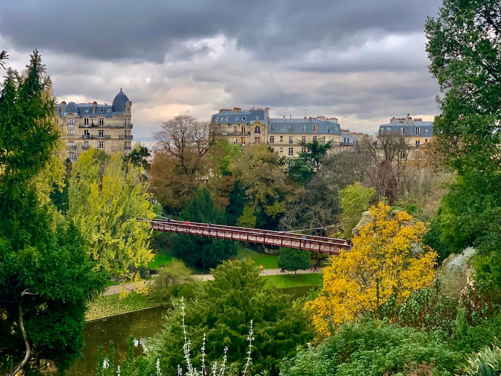 Vista spettacolare del Parc des Buttes-Chaumont con alberi verdissimi, un ponte sospeso rosso e gli edifici parigini classici sotto un cielo nuvoloso.