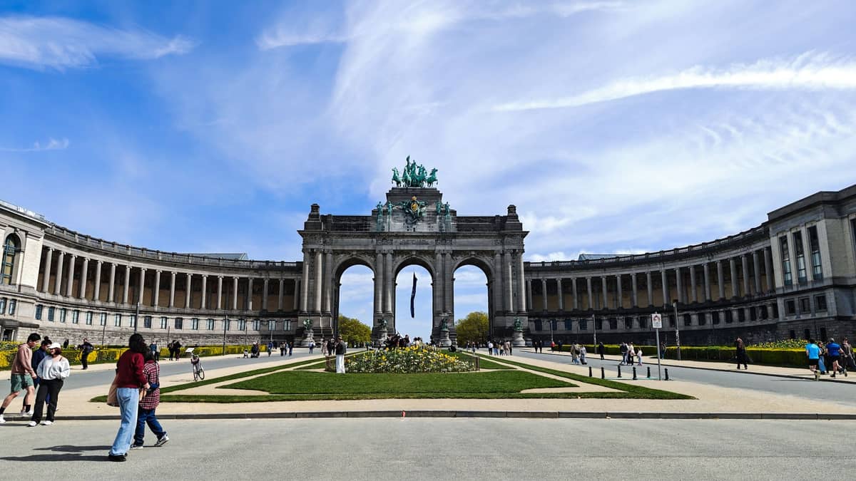 Vista ampla do Parc du Cinquantenaire mostrando o monumental arco do triunfo de três passagens, ladeado por colunatas curvas e pessoas aproveitando o parque num dia ensolarado.