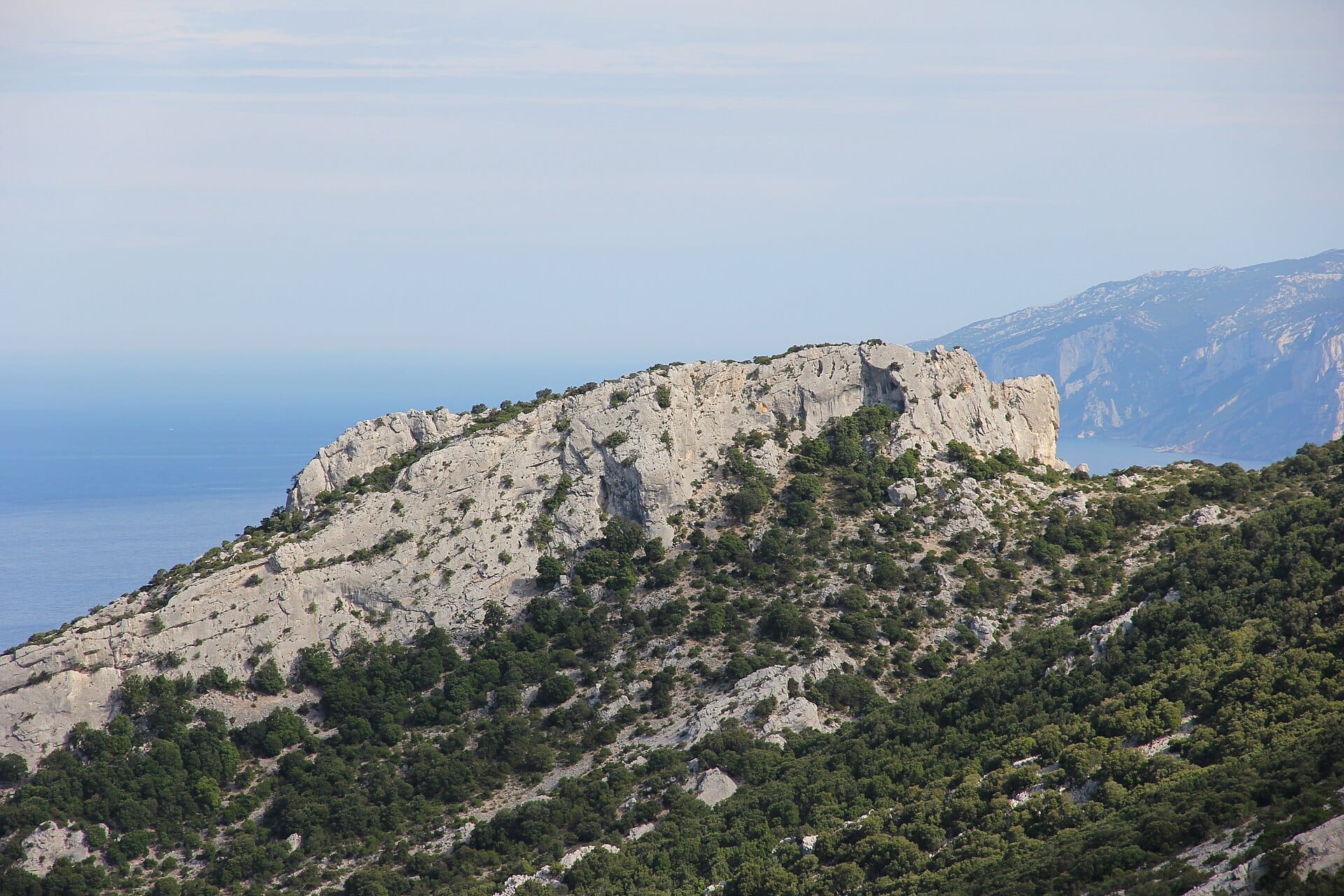 Felsige Kalksteinklippen und grüne Vegetation des Parco Nazionale del Golfo di Orosei e del Gennargentu, mit weiten Blicken auf das blaue Meer und die Berge.