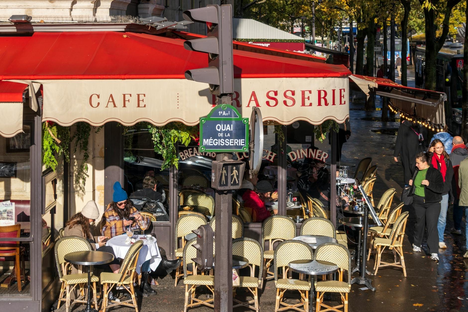 Klassisches Pariser Straßencafé mit roter Markise, Außenbestuhlung und speisenden Gästen an Tischen neben einem baumgesäumten Bürgersteig.