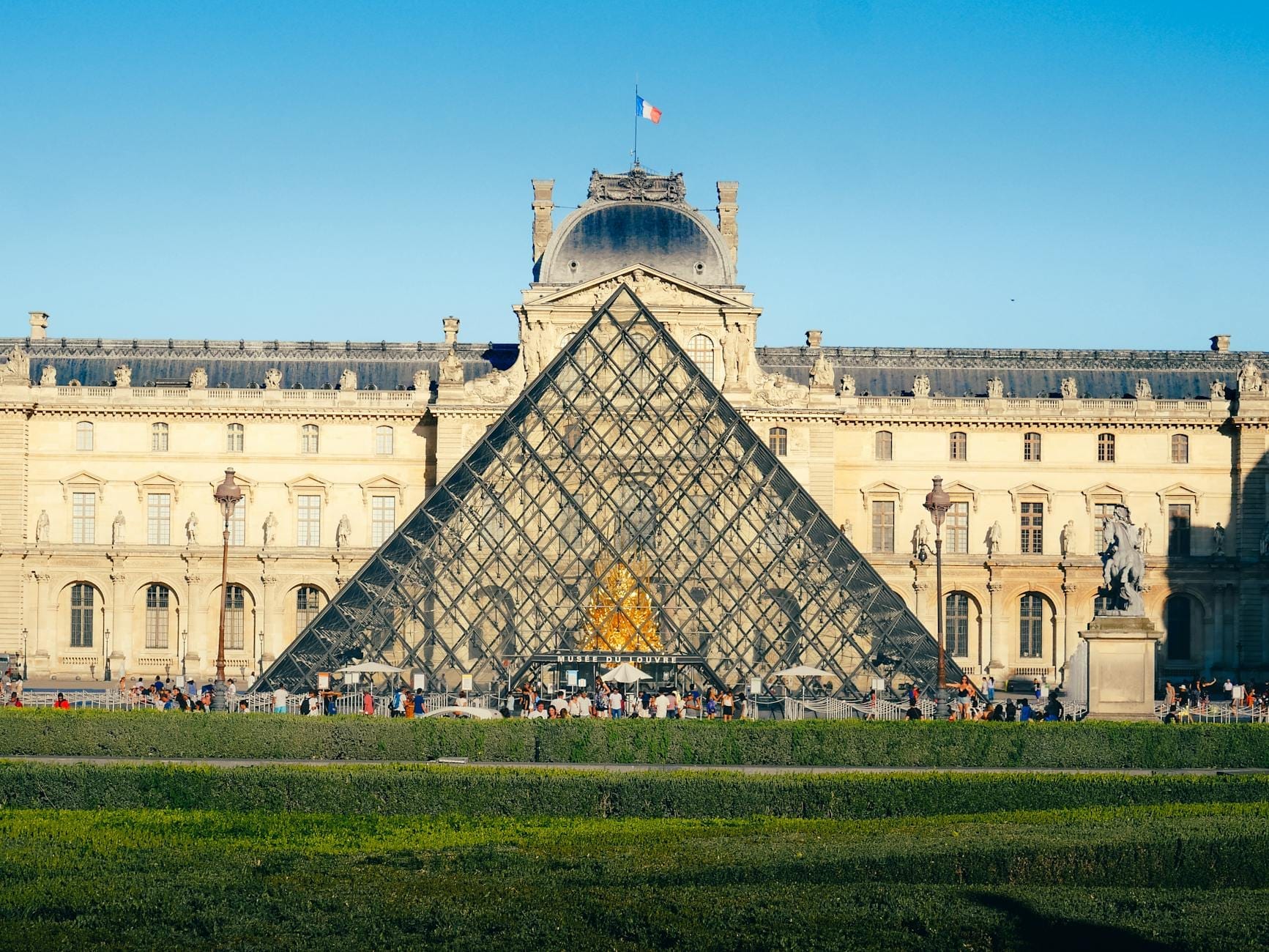 Weitwinkelansicht der Glaspyramide des Louvre und des Museumseingangs mit wartenden Menschen draußen unter einem klaren blauen Himmel.