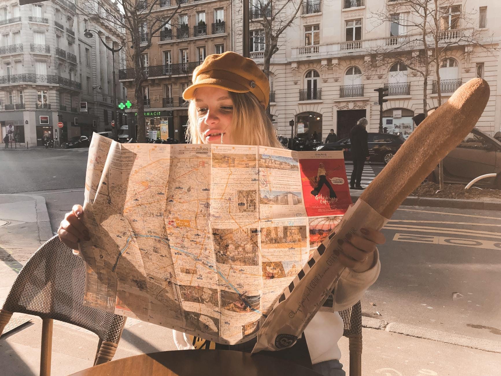 Frau sitzt in einem Pariser Straßencafé, liest einen Stadtplan und hält ein Baguette, im Hintergrund Pariser Gebäude.