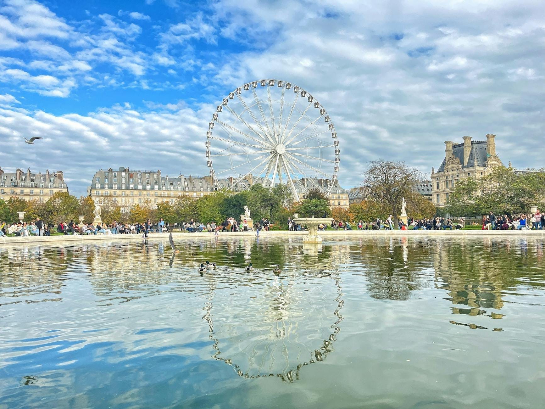 Menschenmassen um den Teich im Jardin des Tuileries in Paris mit Riesenrad und historischen Gebäuden im Hintergrund unter einem teils bewölkten Himmel.