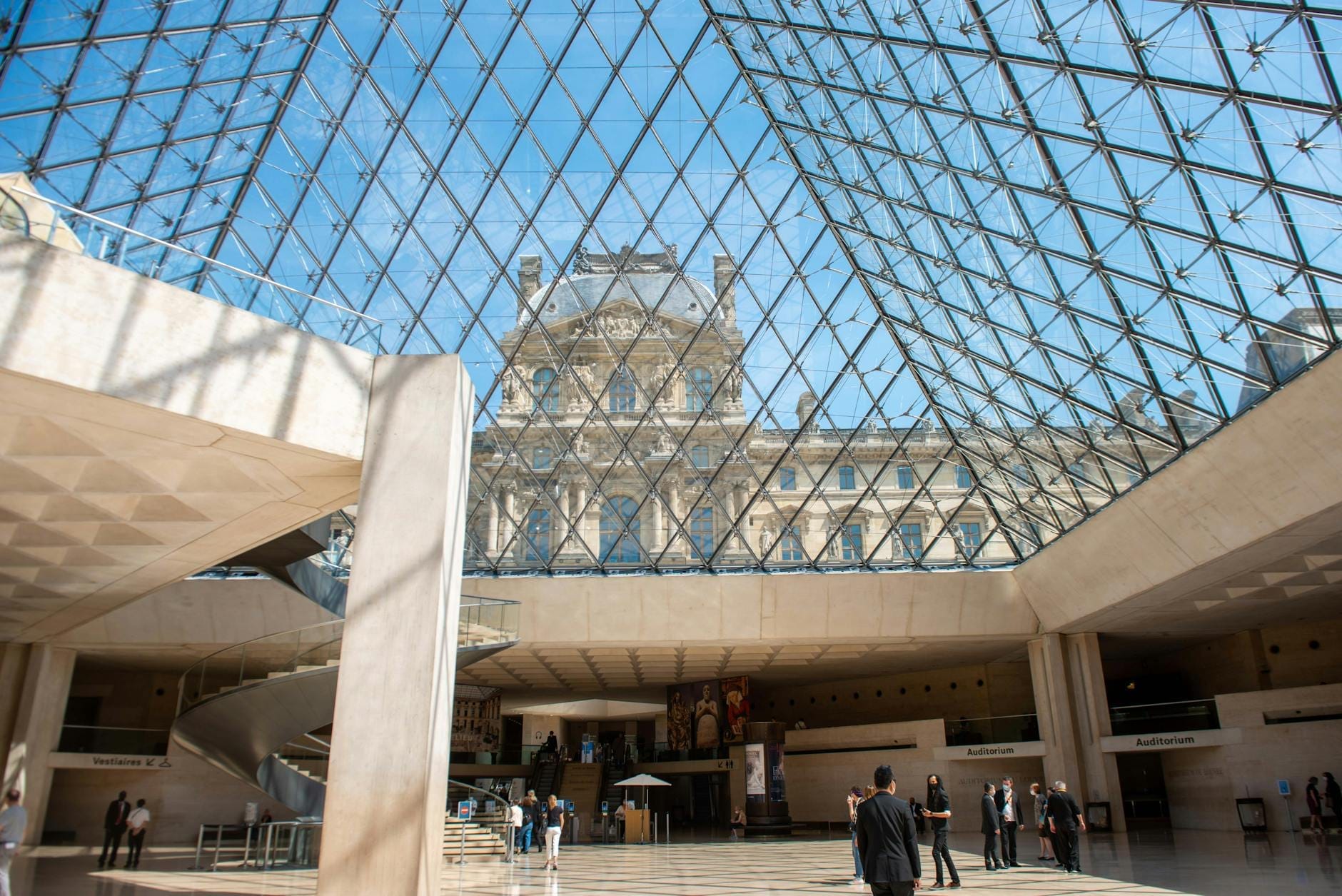 Blick von innen aus dem Louvre-Museum durch die Glaspyramide nach draußen, mit gehenden Menschen und dem historischen Gebäude im Hintergrund.