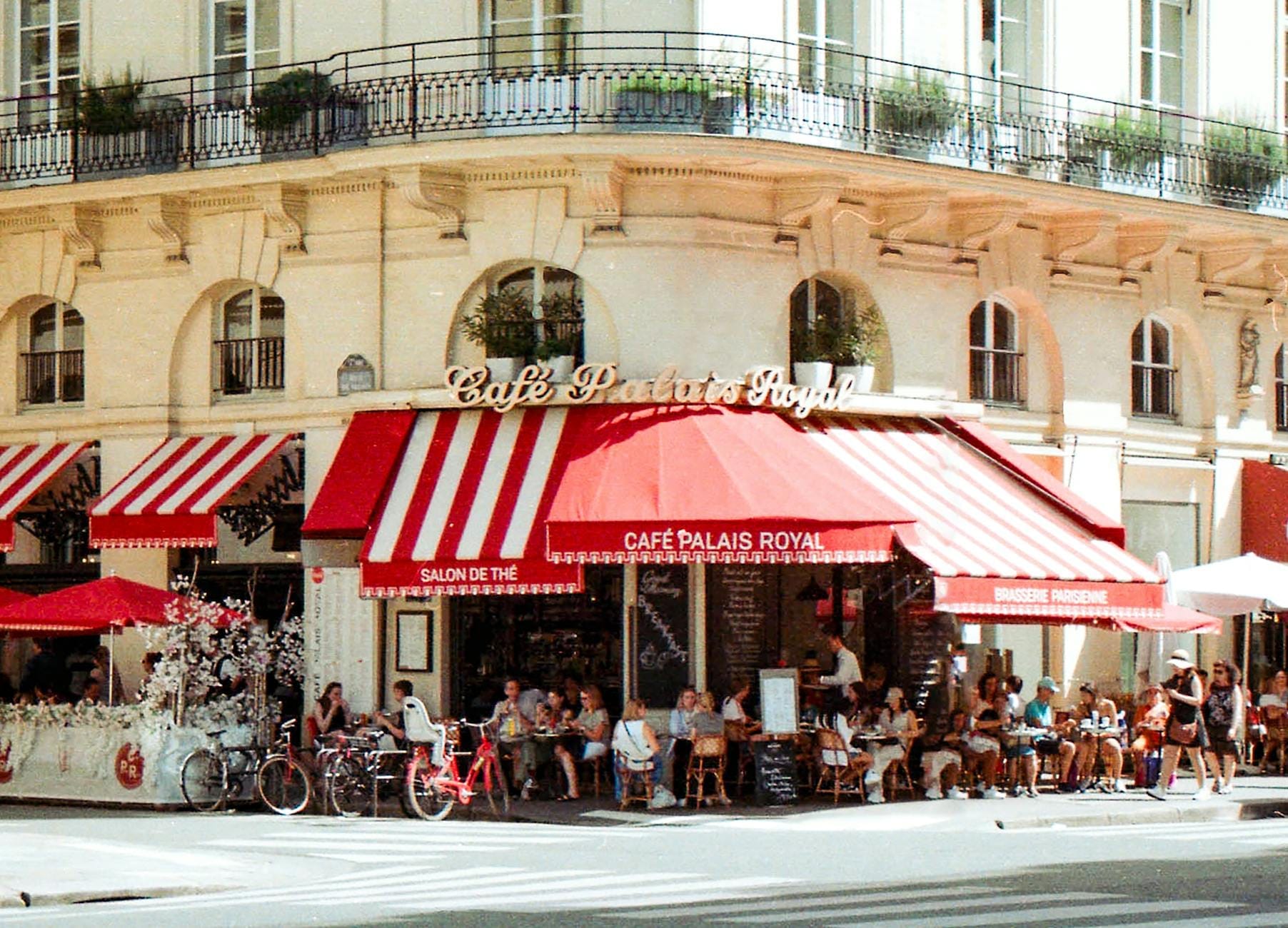 Außentische im Café Palais Royal in Paris, mit speisenden Gästen unter rot gestreiften Markisen an einem sonnigen Tag.