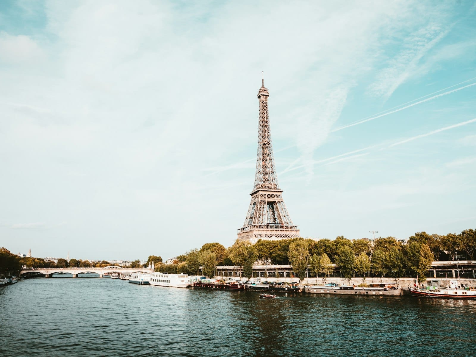 Vista panoramica della Torre Eiffel lungo la Senna in una giornata serena, con barche, alberi e lo skyline di Parigi sullo sfondo.