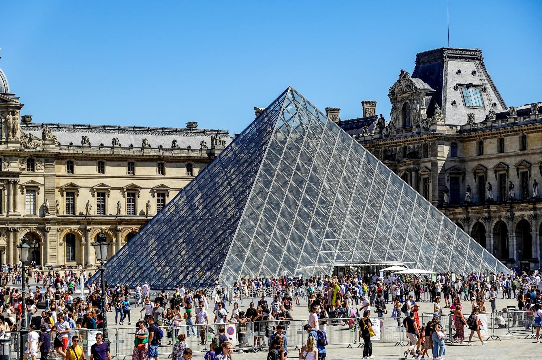 Große Menschenmenge vor dem gläsernen Pyramideneingang des Louvre an einem sonnigen Tag, die die typisch langen Warteschlangen im Sommer veranschaulicht.