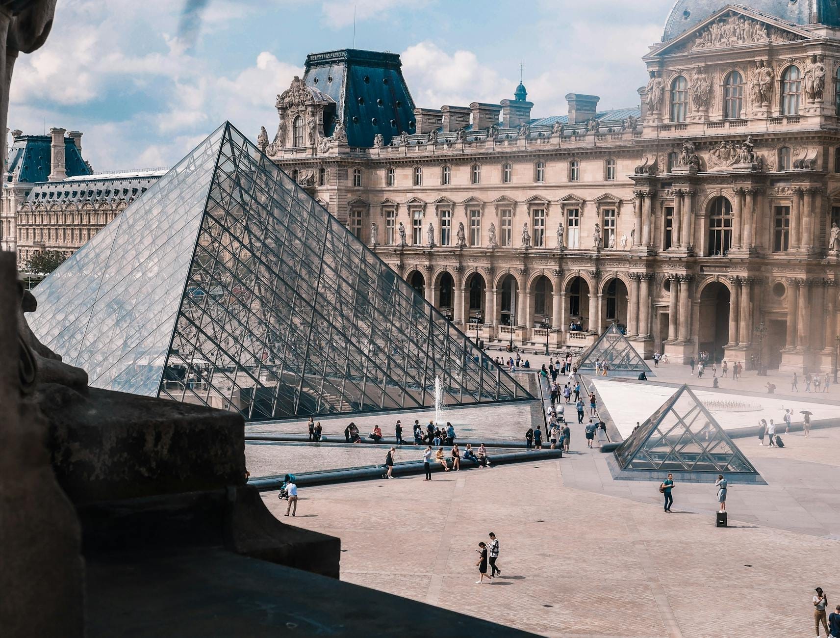 Foto des Innenhofs des Musée du Louvre mit der ikonischen Glaspyramide und den umliegenden historischen Gebäuden sowie Besuchern, die über den Platz spazieren.