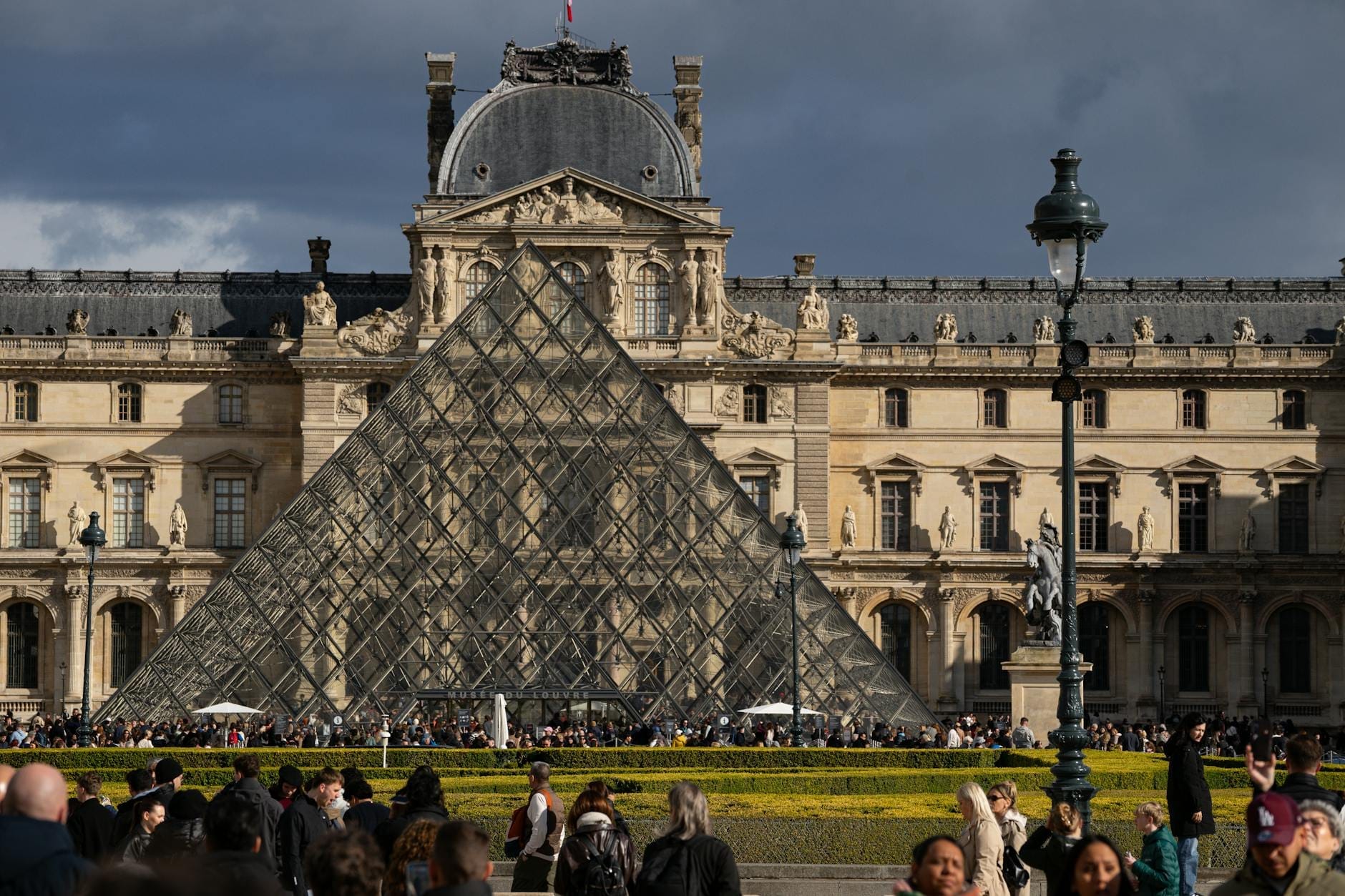 Il Museo del Louvre e la sua iconica entrata a piramide di vetro a Parigi, con molti visitatori in primo piano sotto un cielo spettacolare.