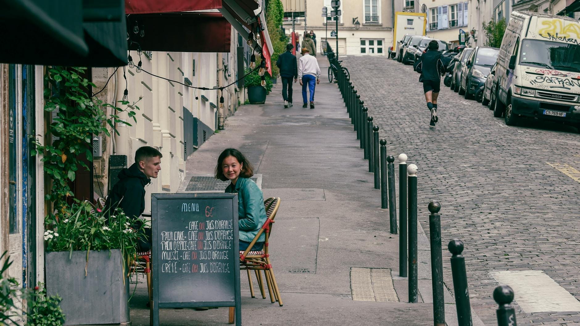 Scena di strada parigina con persone che pranzano in un piccolo caffè all'aperto, lavagna del menù con i prezzi davanti e la tipica architettura di Parigi sullo sfondo.