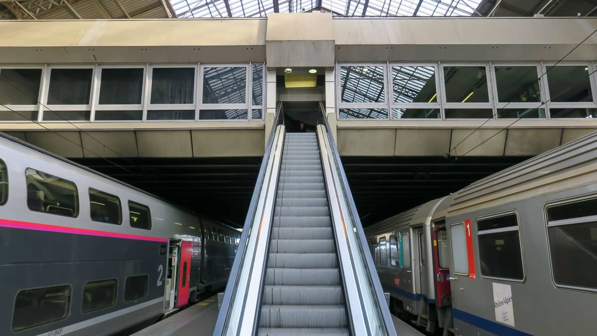 Escada rolante entre dois trens internacionais em uma estação ferroviária moderna com teto de vidro, com uma atmosfera prática e funcional.