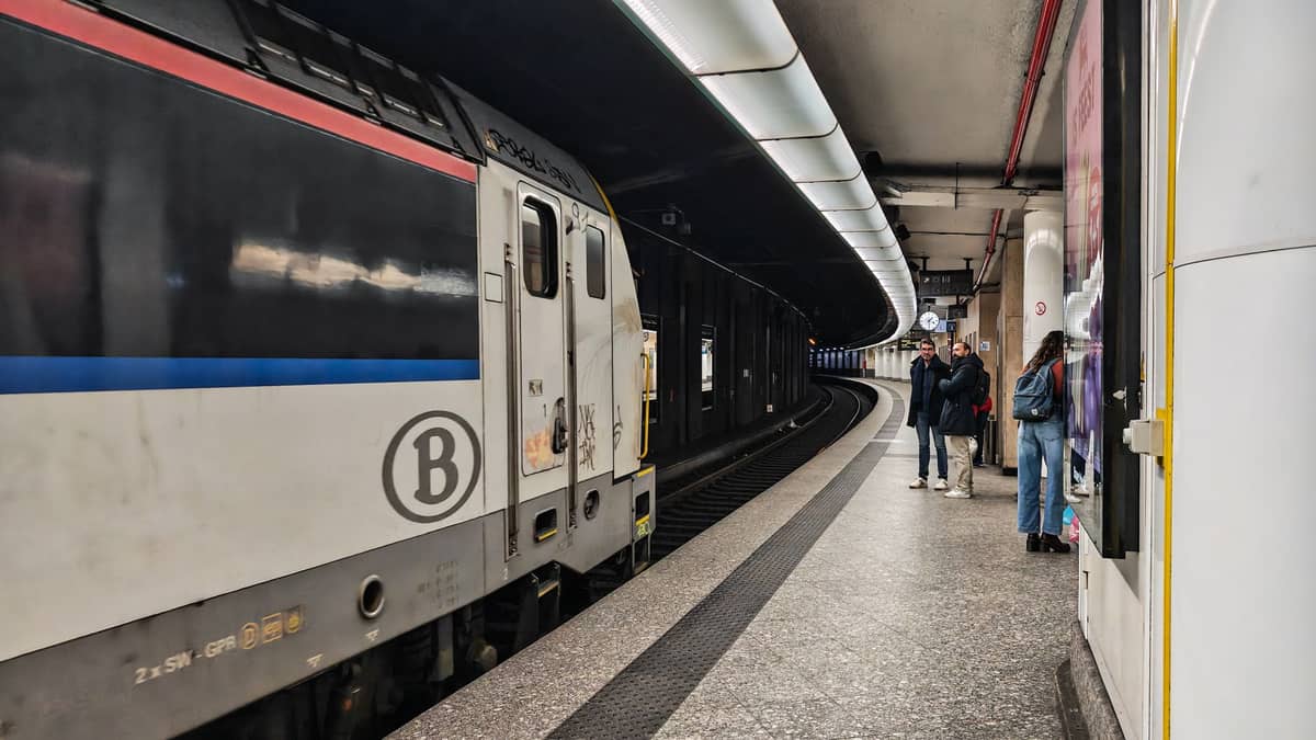 Um moderno trem belga da SNCB parado em uma plataforma de uma estação em Bruxelas enquanto passageiros aguardam na plataforma de azulejos sob uma iluminação curva.