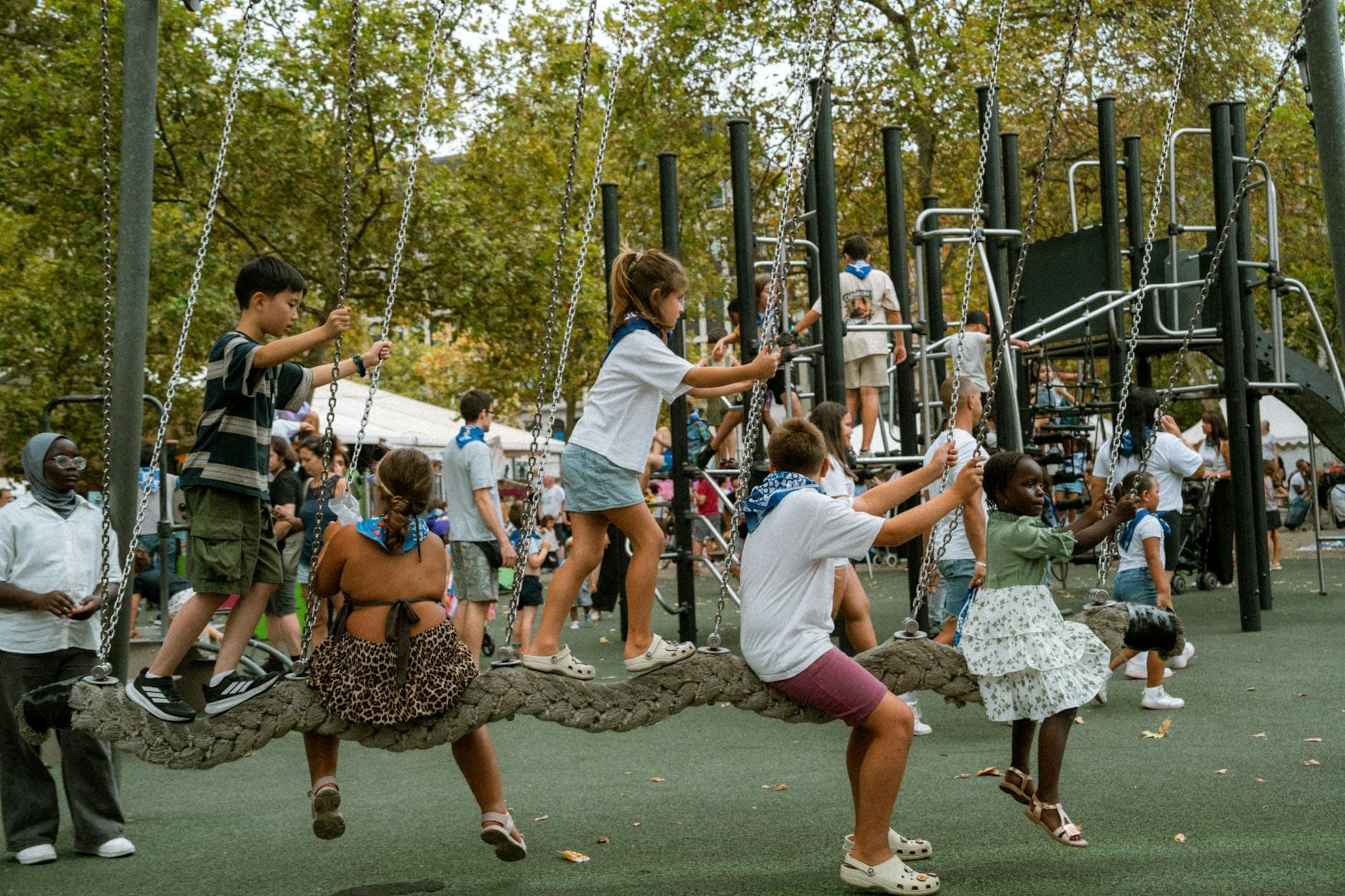 Gruppo di bambini che giocano energicamente insieme su moderne attrezzature da gioco in un parco parigino con adulti e alberi sullo sfondo.