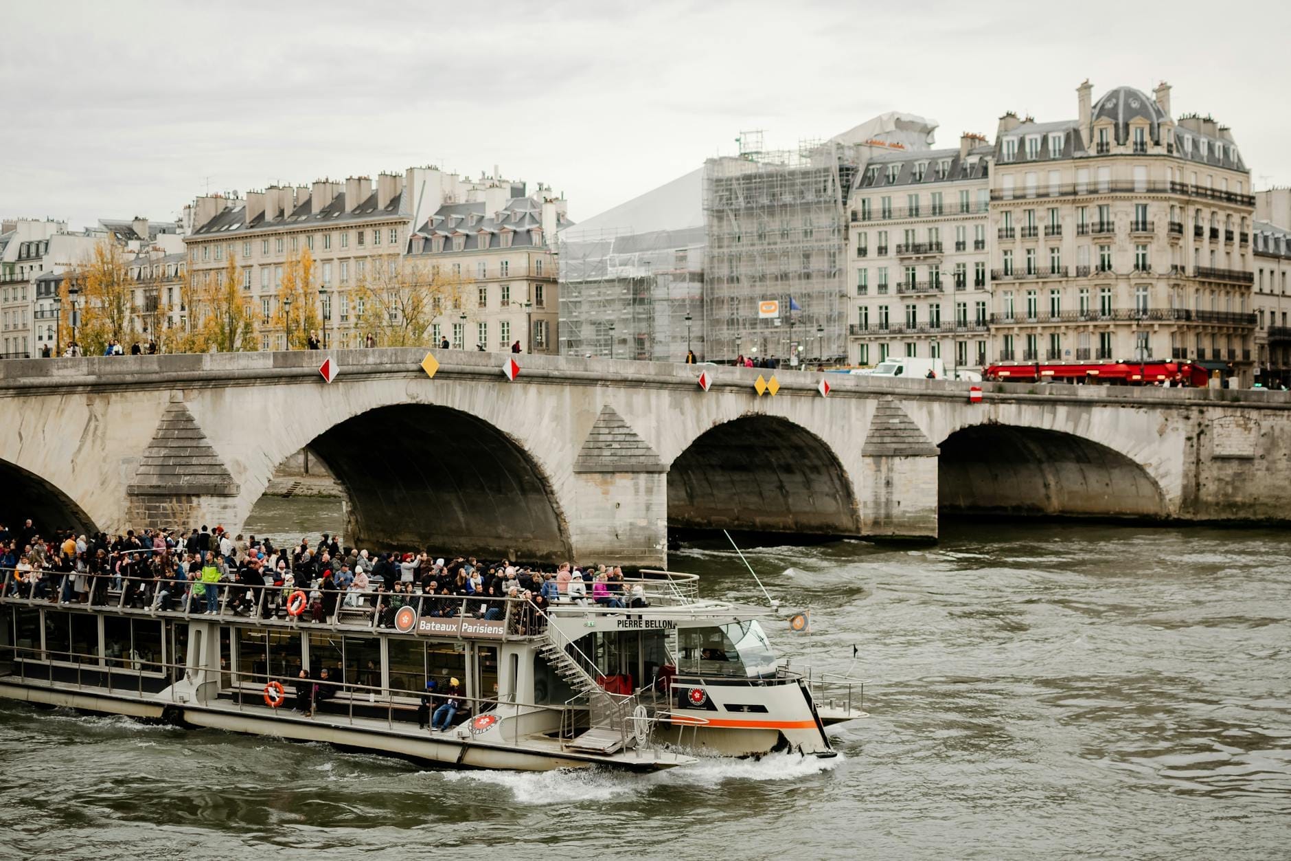 Un lungo battello turistico pieno di persone passa sotto uno storico ponte in pietra sulla Senna a Parigi, con edifici parigini sullo sfondo.
