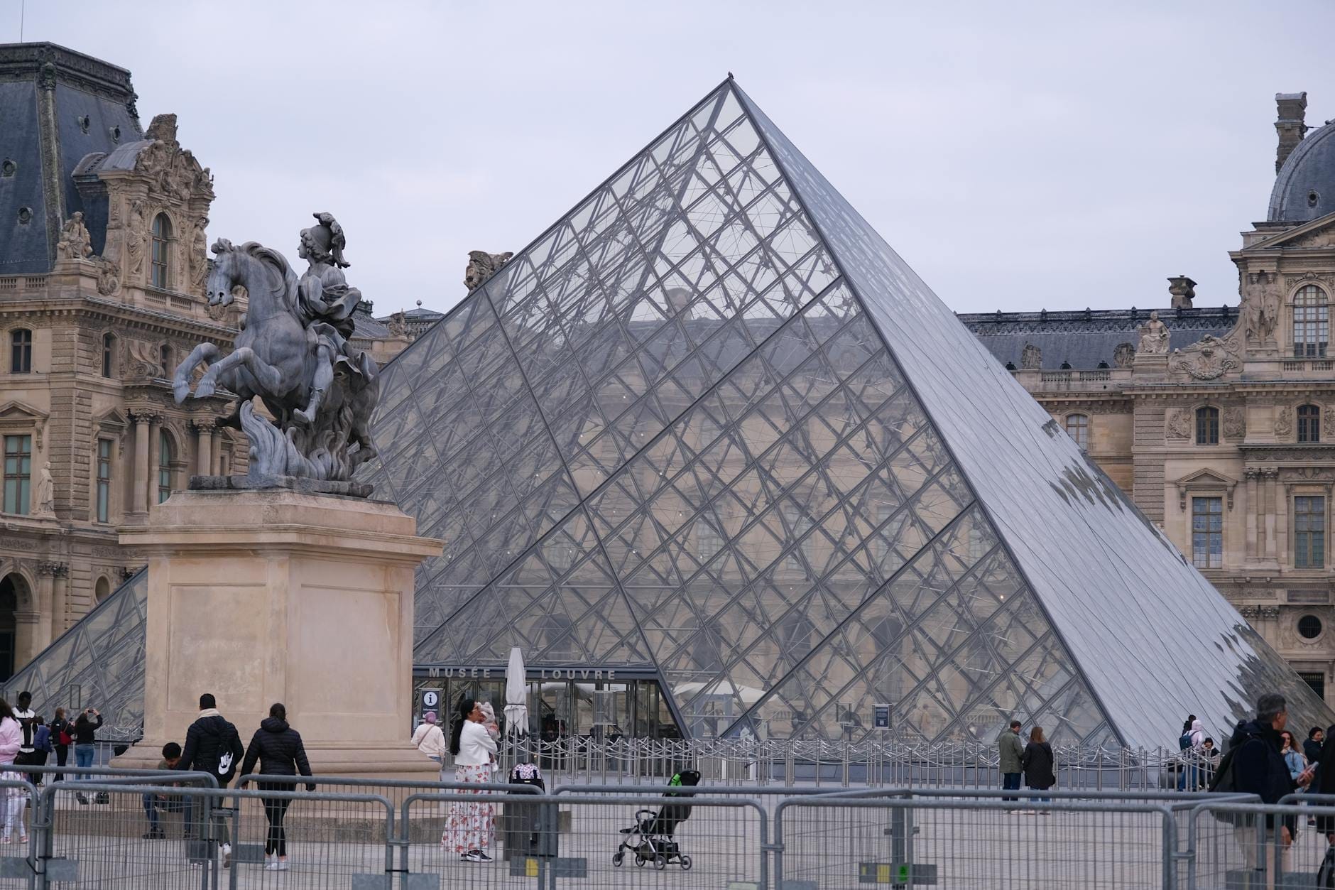 Vista dell'ingresso della Piramide del Louvre a Parigi con famiglie, una statua in primo piano e persone che camminano per la piazza.