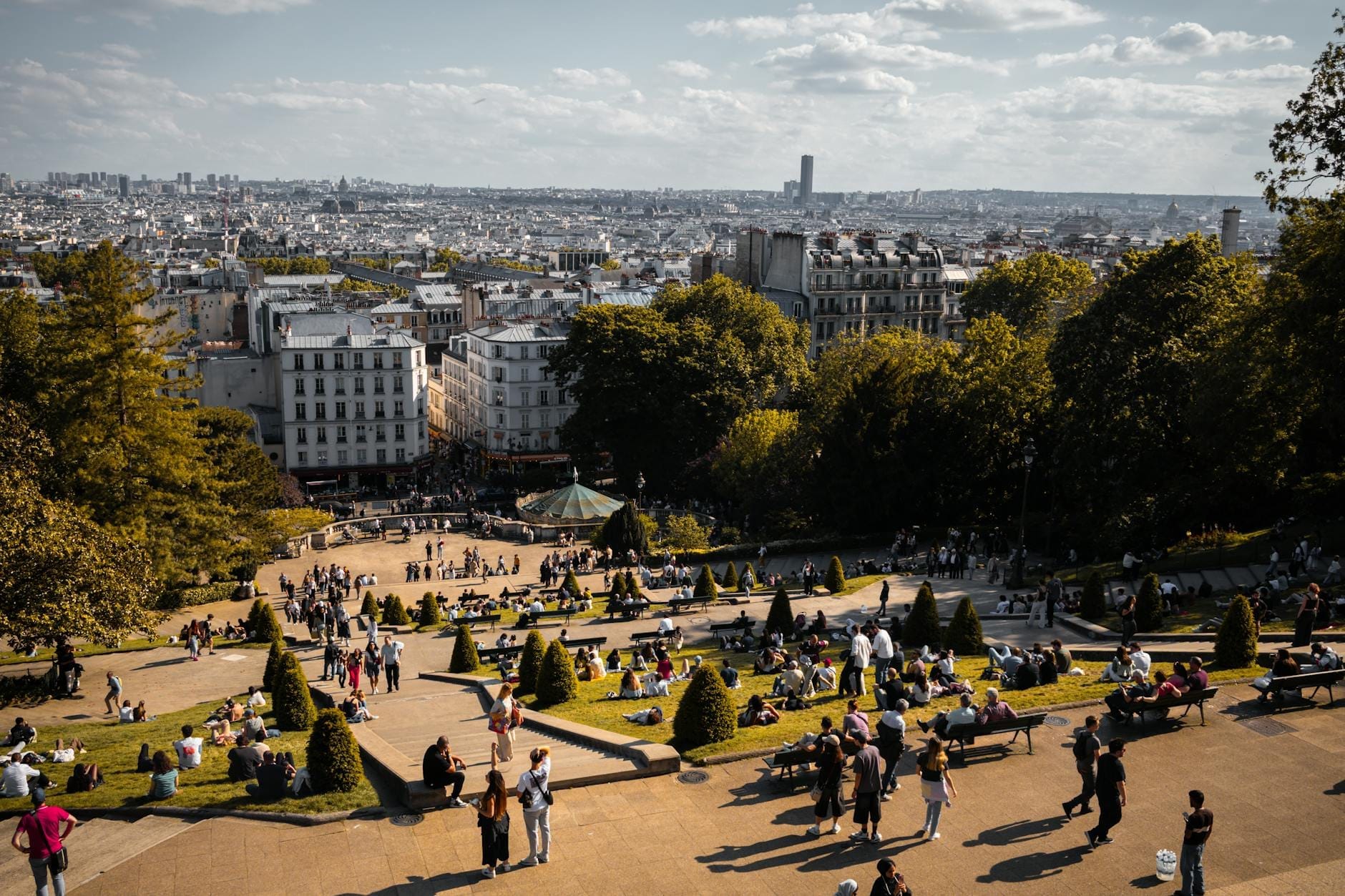 Vista su un vivace parco parigino pieno di famiglie e bambini, con panchine, alberi e classici edifici cittadini sullo sfondo sotto un cielo soleggiato.