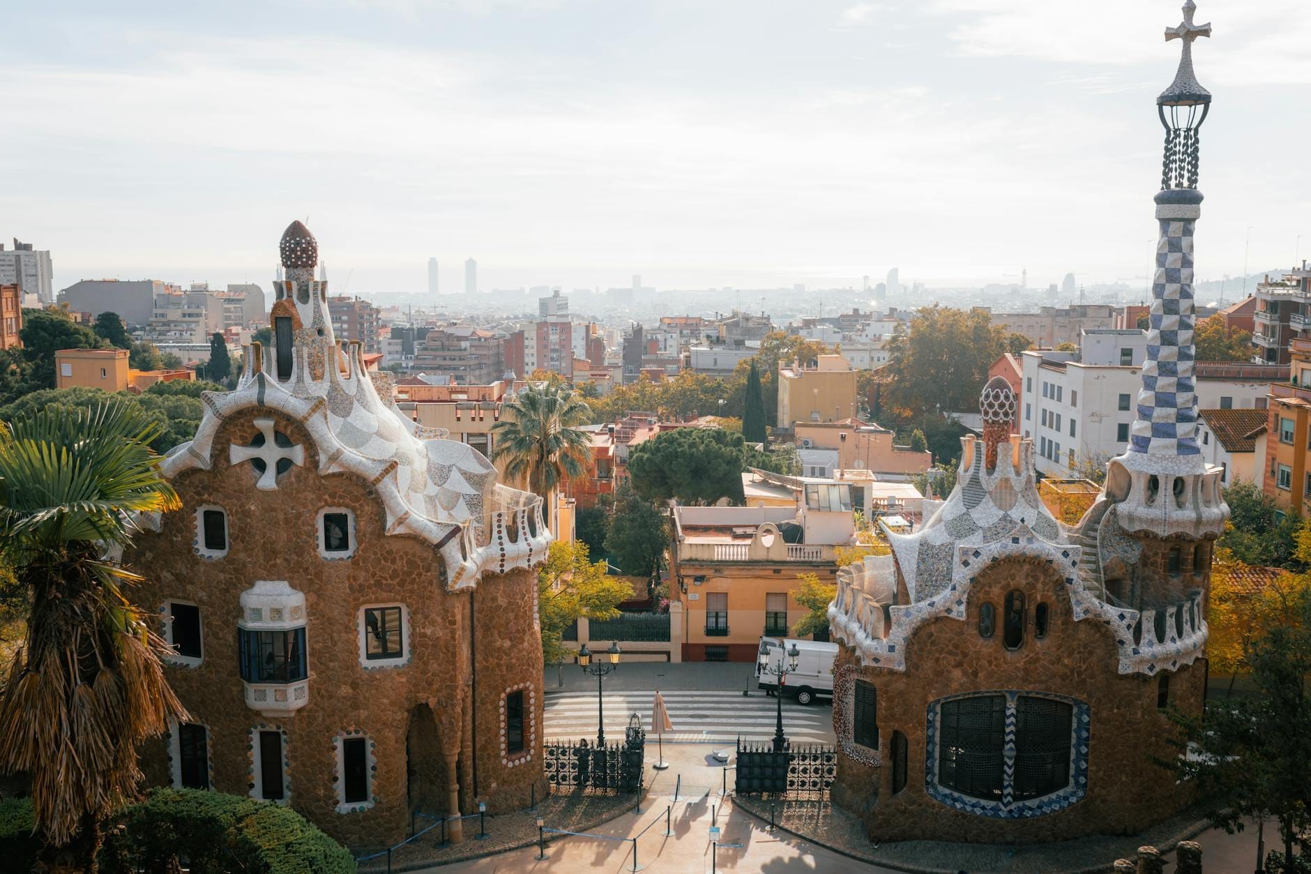 Wide landscape view from Park Güell’s entrance, featuring Gaudí’s whimsical gatehouses and Barcelona’s skyline stretching toward the Mediterranean on a bright sunny day.