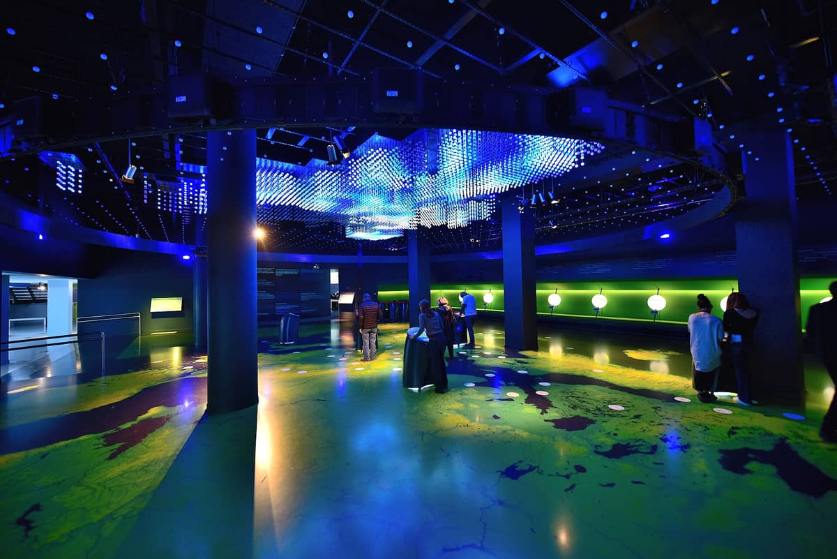 Interior do centro de visitantes Parlamentarium, com exposições interativas e um mapa iluminado sob luzes azuis, enquanto visitantes exploram o espaço moderno e imersivo.