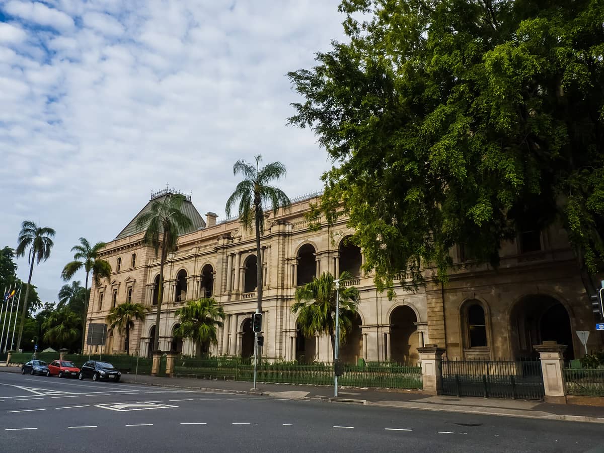Szeroki widok na Queensland Parliament House w Brisbane – imponująca kolonialna architektura, łukowe okna, palmy i żelazne ogrodzenie pod częściowo zachmurzonym niebem.