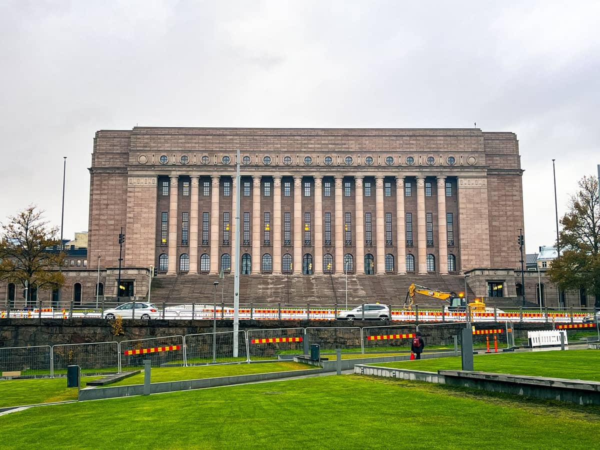 Vista frontal do Parlamento de Helsinque com sua imponente fachada de pedra, fileiras de altas colunas coríntias e um amplo gramado verde em primeiro plano.