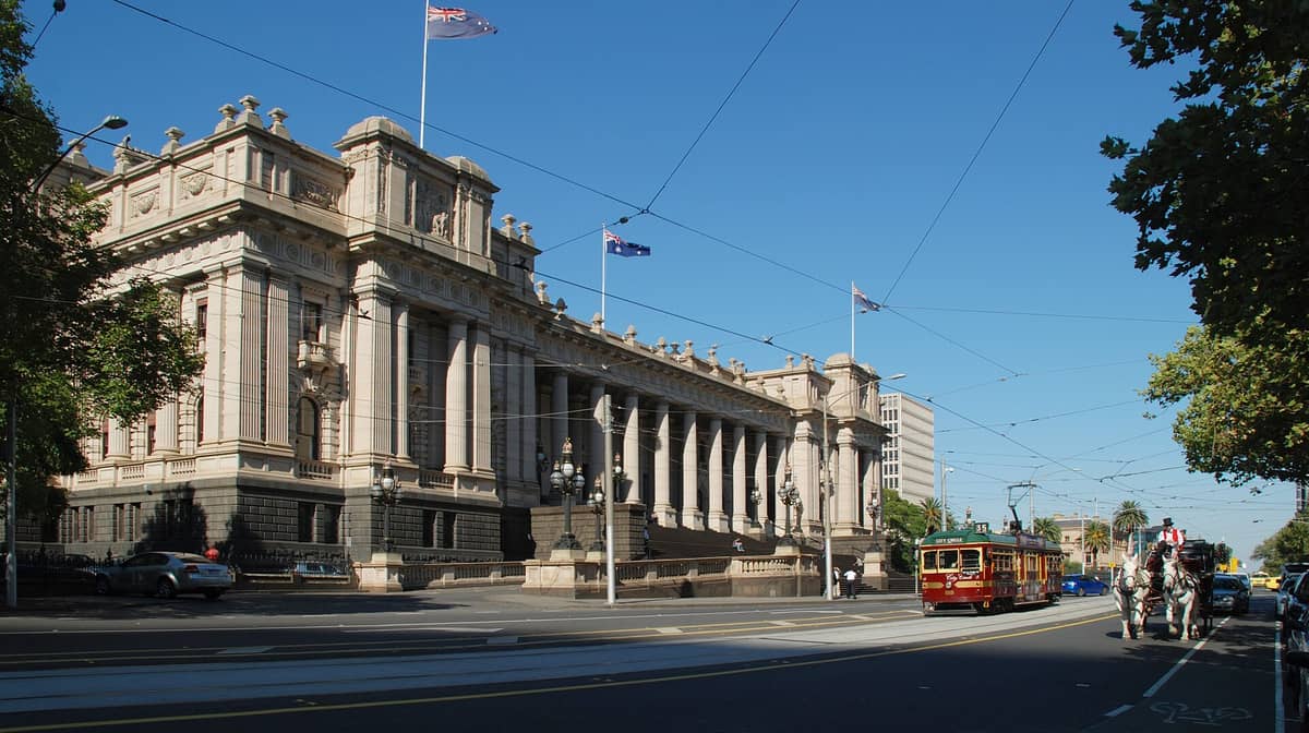 Weiter Blick auf die prächtige neoklassizistische Fassade des Parliament House Melbourne an der Spring Street im Sonnenlicht, mit einer vorbeifahrenden Tram, Straßenbäumen und Menschen davor.