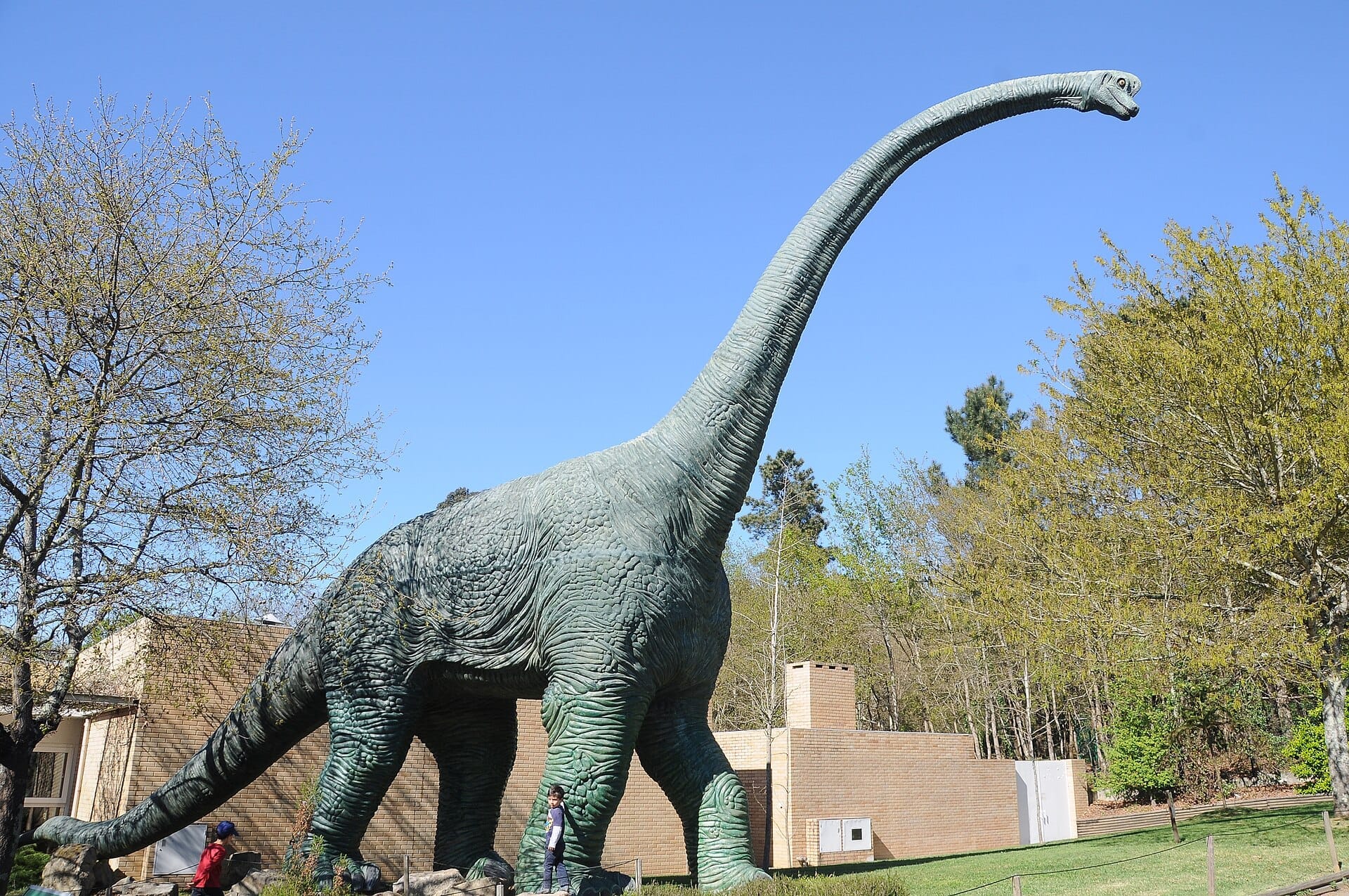 Grande statua di dinosauro al Parque Biológico de Gaia circondata da alberi primaverili ed edifici in mattoni sotto un cielo azzurro limpido.