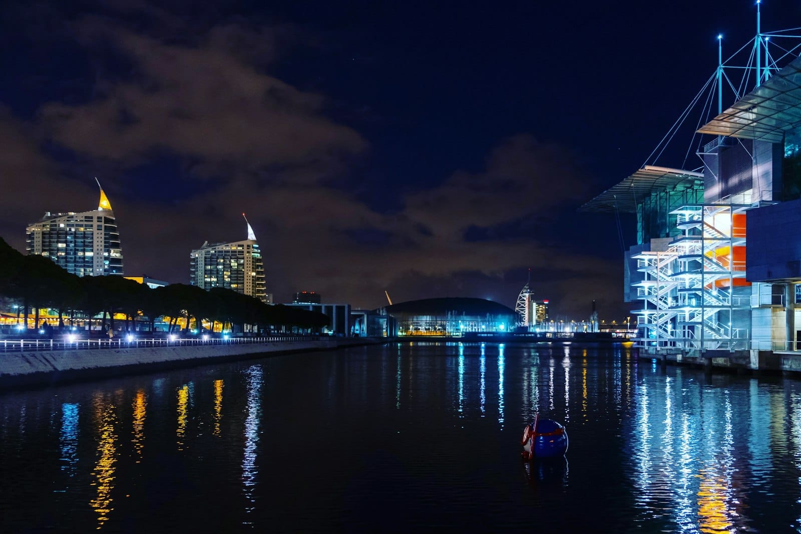 วิวริมน้ำยามค่ำคืนของ Parque das Nações กับตึกสมัยใหม่ หอคอยสัญลักษณ์ และ Oceanário de Lisboa ที่สะท้อนแสงในแม่น้ำใต้ท้องฟ้าอันน่าตื่นตา