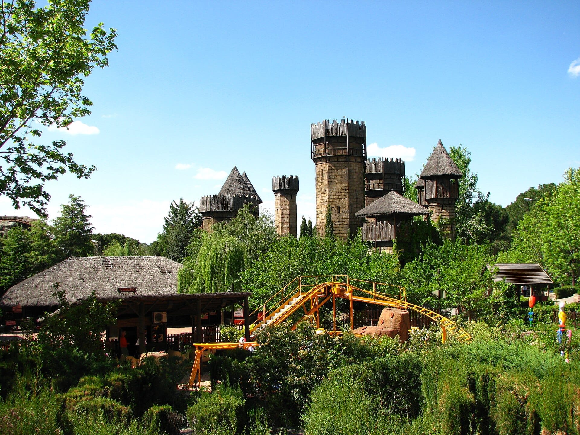 Torri di un castello in pietra e un ottovolante giallo circondati da rigogliosa vegetazione al Parque de Atracciones de Madrid sotto un cielo azzurro limpido.