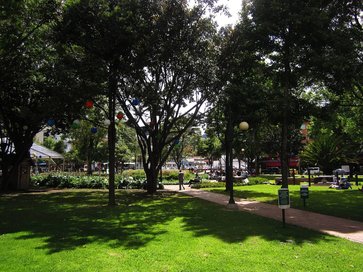 Shady green parkland in Parque de la 93, Bogotá, with trees, lawns, walking paths, people relaxing, and nearby restaurants visible on a sunny afternoon.