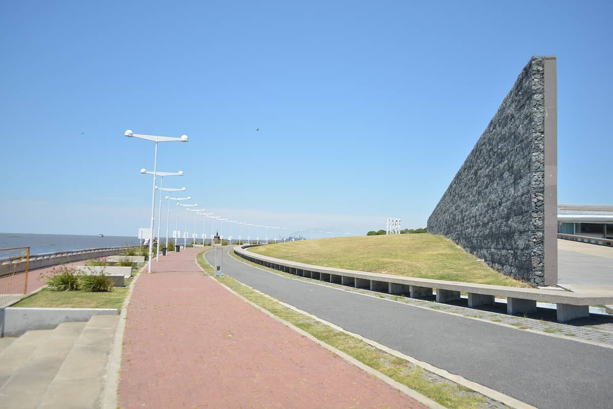 มุมกว้าง Parque de la Memoria ในบัวโนสไอเรส ให้เห็นกำแพงรำลึก ทางเดินโค้งเลียบแม่น้ำ และสนามหญ้าใต้ท้องฟ้าใส
