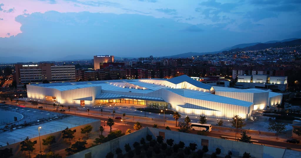 มุมมองทางอากาศของ Parque de las Ciencias Granada ยามพลบค่ำ พร้อมอาคารกระจกสมัยใหม่ที่สว่างไสวและแสงไฟของเมืองในพื้นหลัง