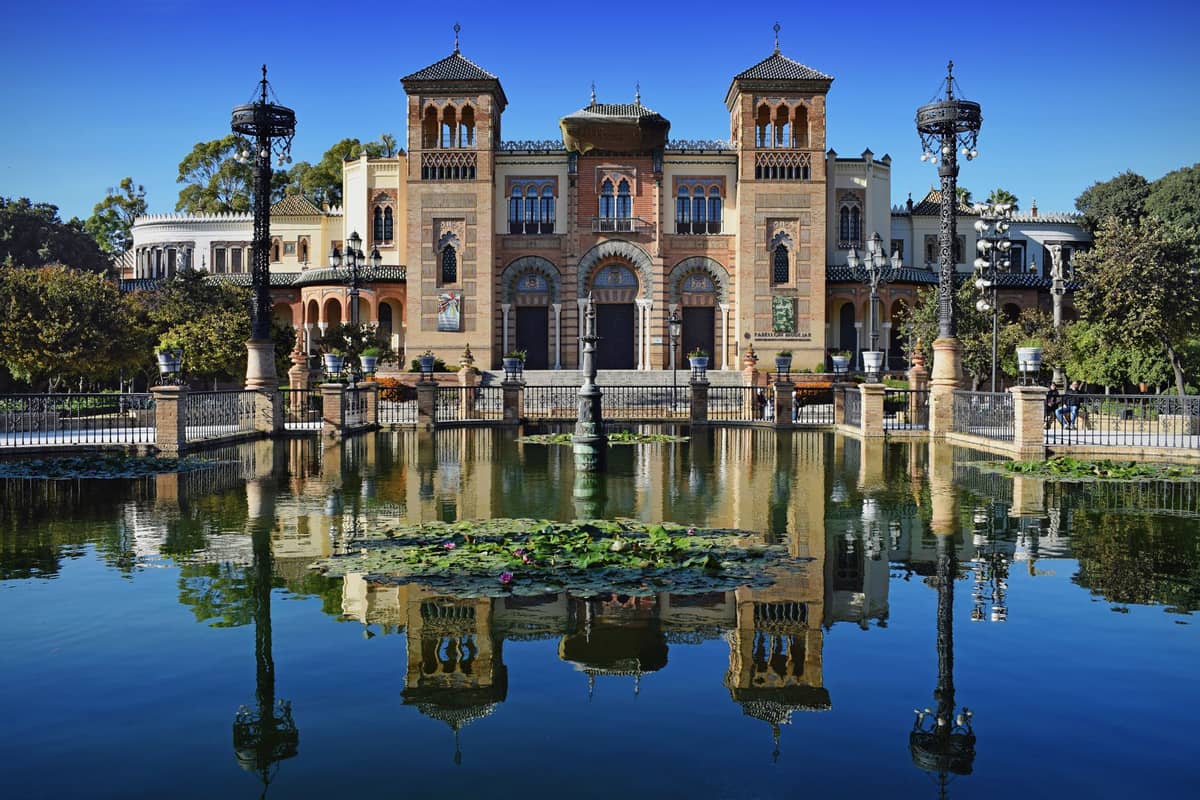 ศาลา Mudejar อันยิ่งใหญ่สะท้อนบนสระน้ำประดับขนาดใหญ่ รายล้อมด้วยสวนร่มรื่นและท้องฟ้าสีฟ้าในสวน Parque de María Luisa ที่เซบีญ่า
