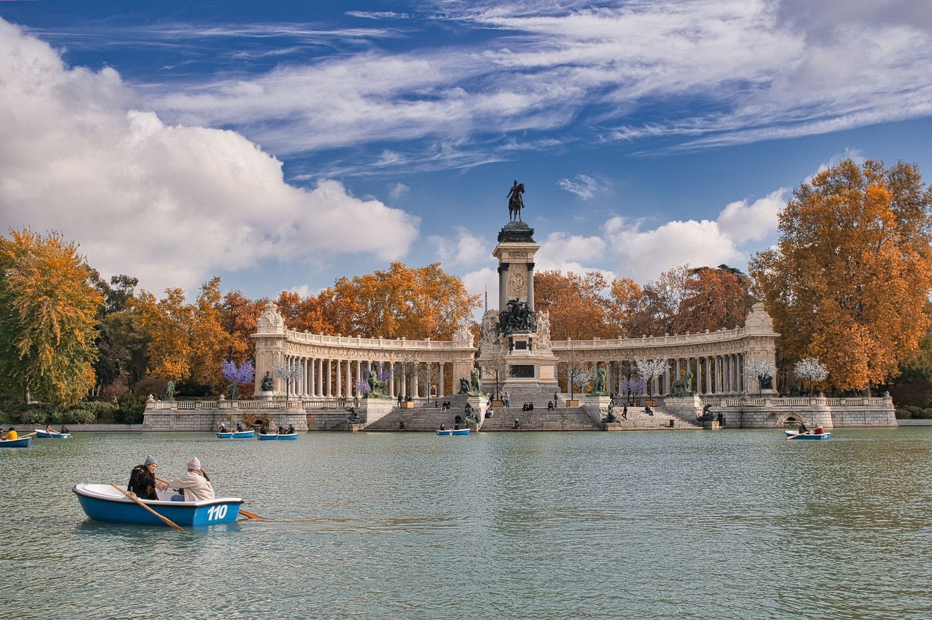 Vista del Monumento ad Alfonso XII con alberi autunnali e cielo azzurro, persone in barca sul grande lago del Parque del Retiro di Madrid.