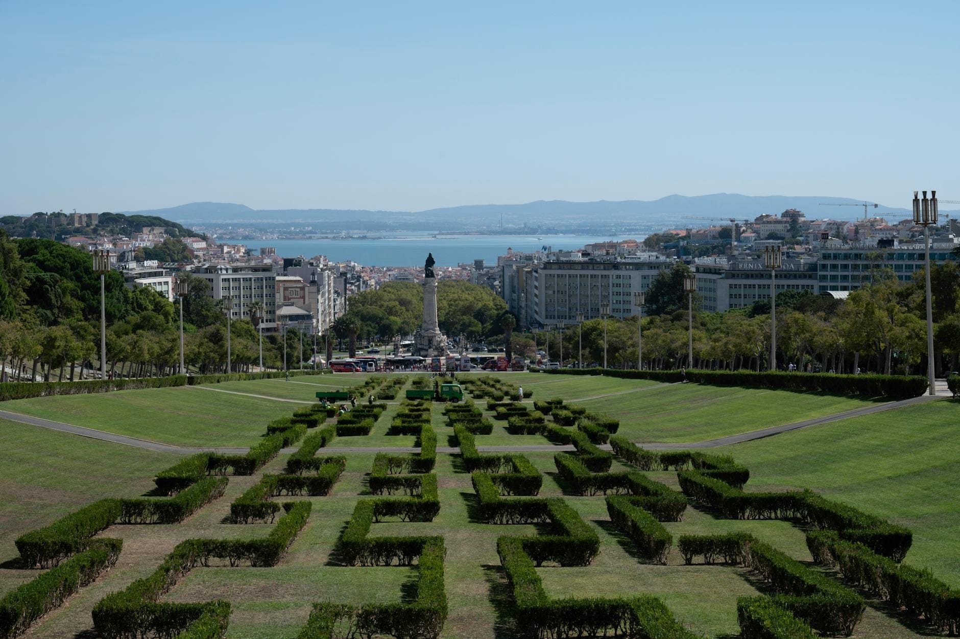 Vista sulle siepi geometriche del Parque Eduardo VII, sul viale centrale e sullo skyline di Lisbona con il fiume Tago e le colline sullo sfondo sotto un cielo blu limpido.