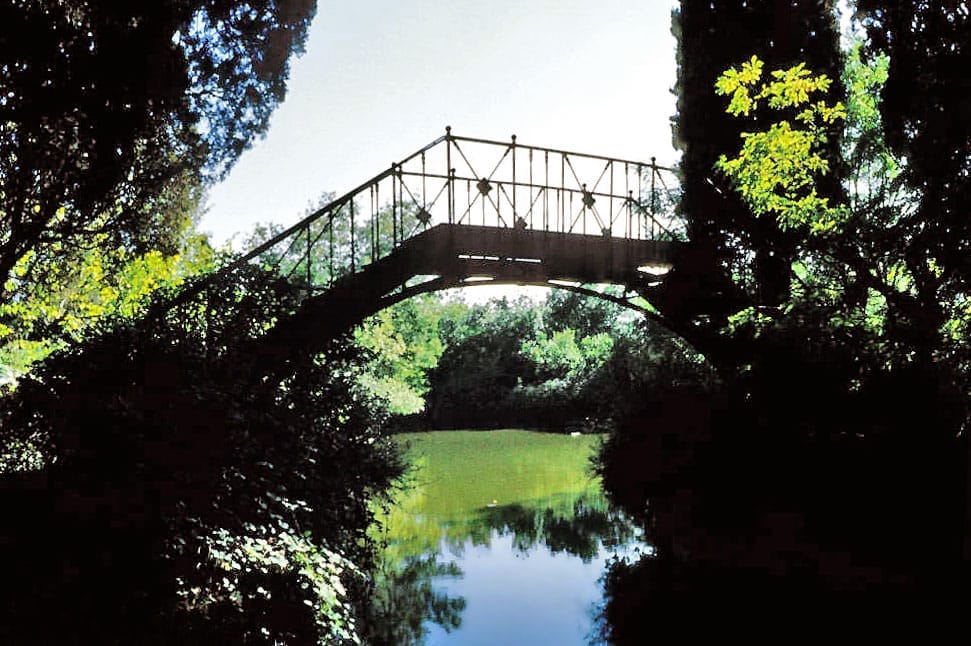 Un ponte in ferro ad arco attraversa uno stagno tranquillo, circondato da una rigogliosa vegetazione nel Parque El Capricho a Madrid.