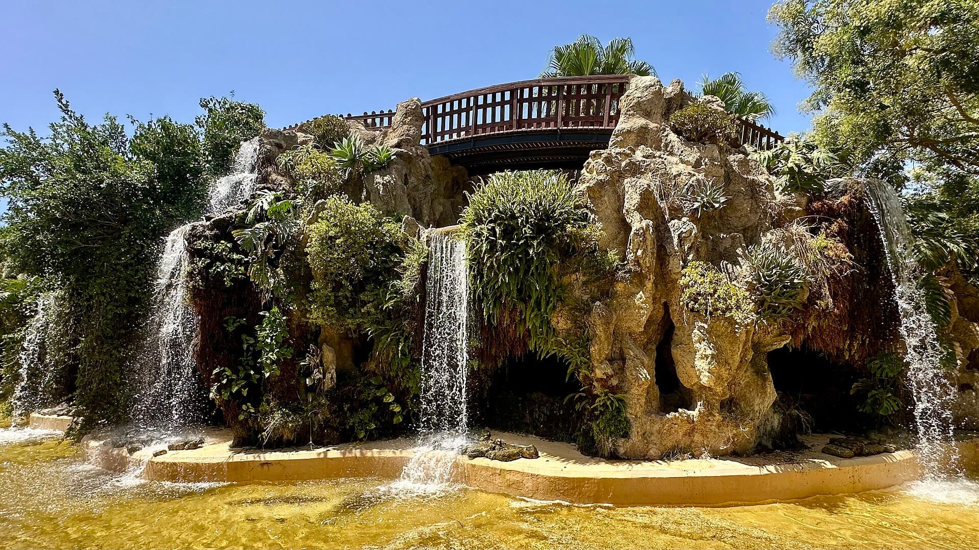 Sunlit view of Parque Genovés waterfall with lush greenery, rock formations, and wooden bridge set against a bright blue sky in Cádiz.