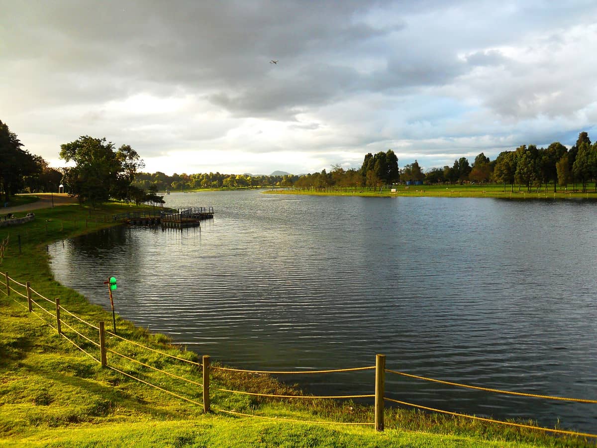 Wide view of the central lake at Parque Metropolitano Simón Bolívar in Bogotá, with grassy banks, calm water, and trees under a dramatic sky.