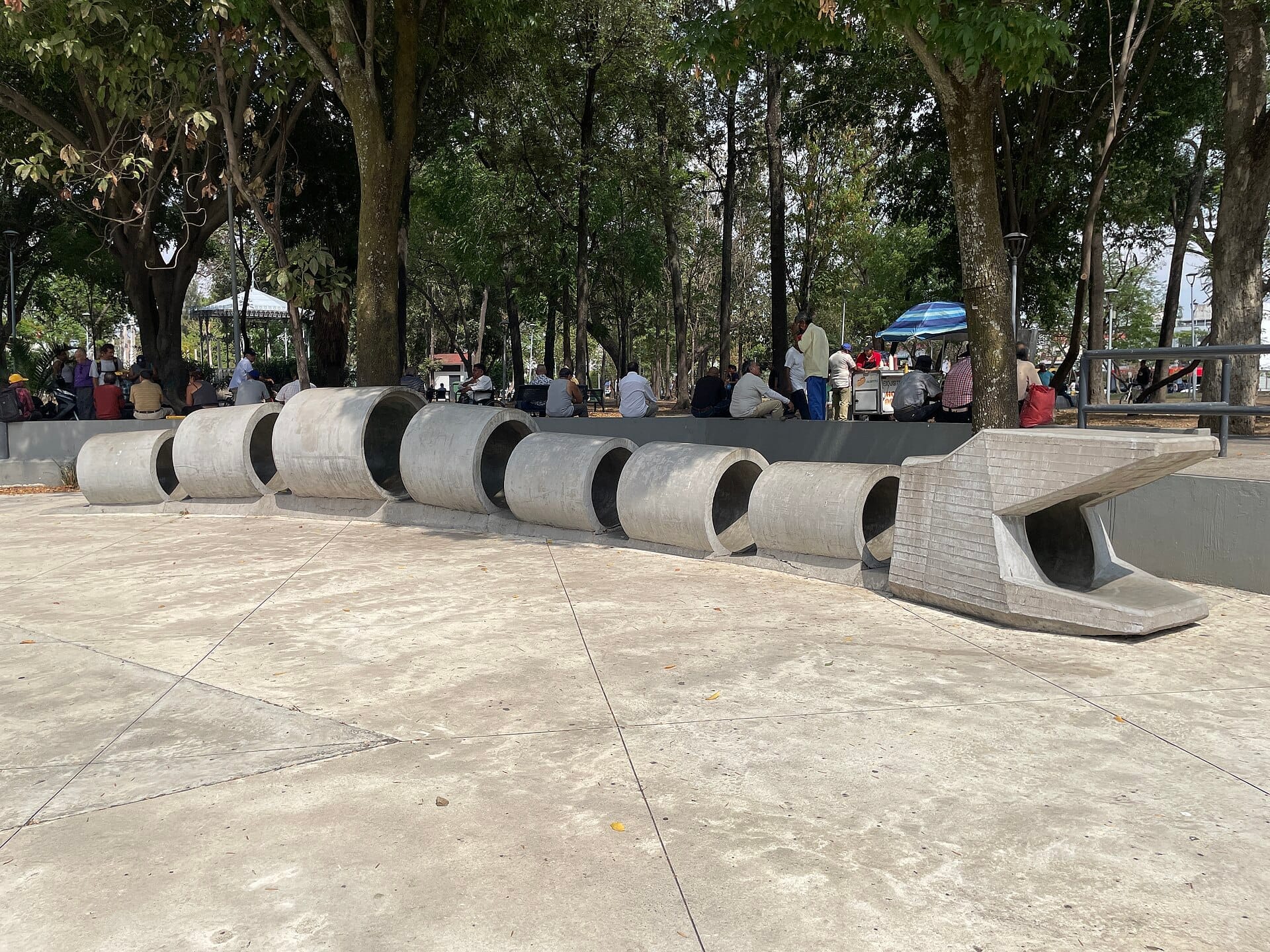 Una scultura in cemento che ricorda un animale segmentato si estende sulla piazza del Parque Morelos, circondata da alberi e aree ombreggiate con sedute e visitatori.
