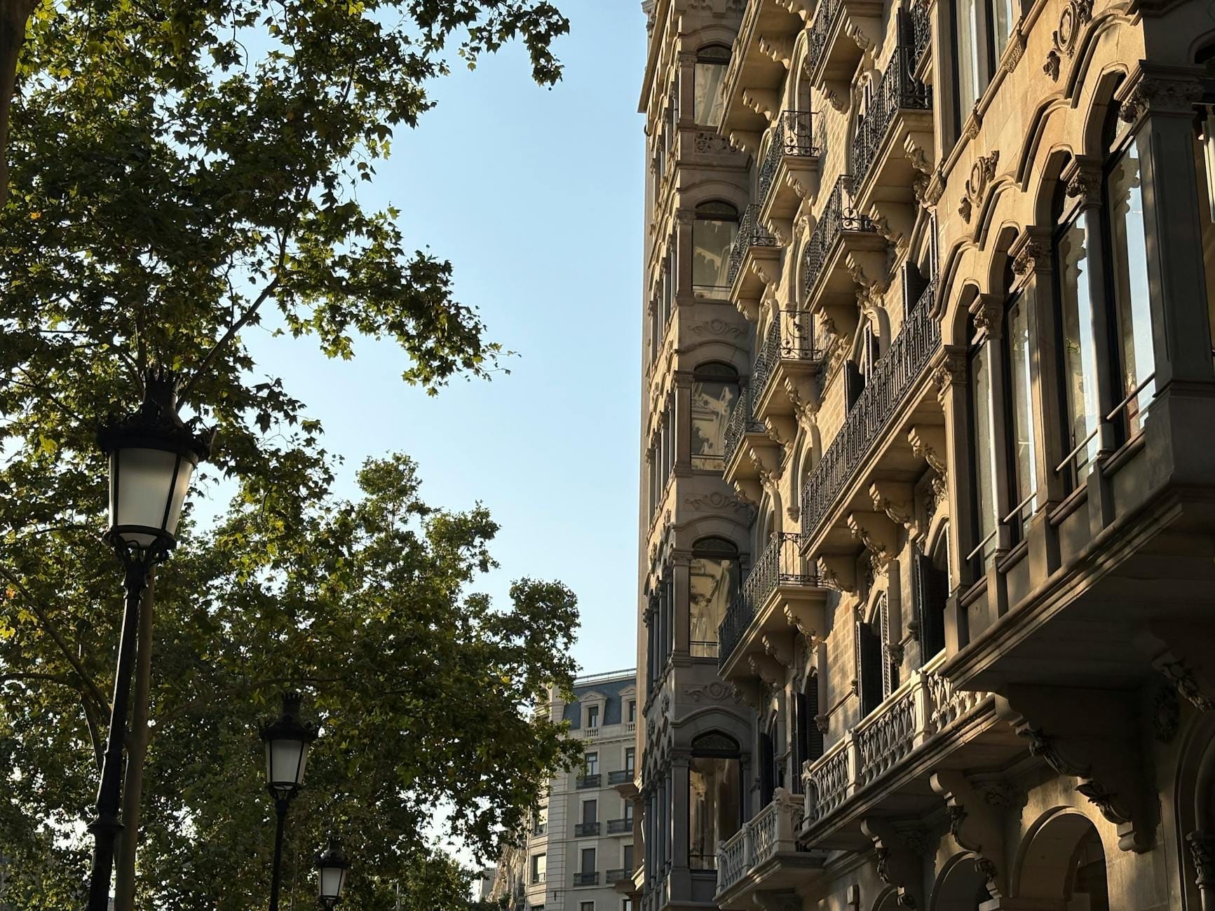 Elegant historic buildings and ornate balconies line Passeig de Gràcia in Barcelona, with lush green trees and vintage street lamps under a clear sky.