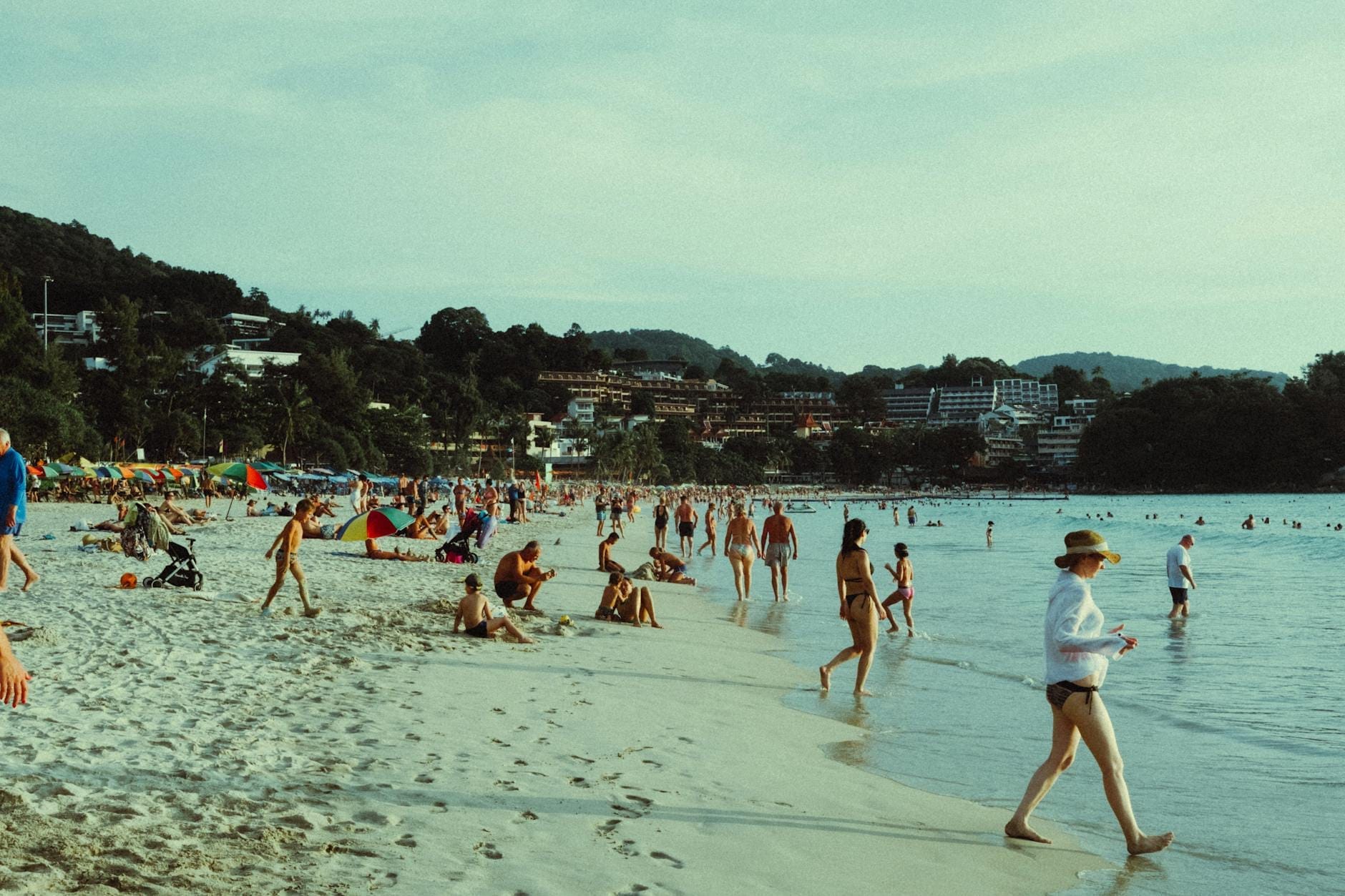 Crowds of tourists relaxing, swimming, and walking along the sandy shoreline of Patong Beach with hotels and greenery in the background.
