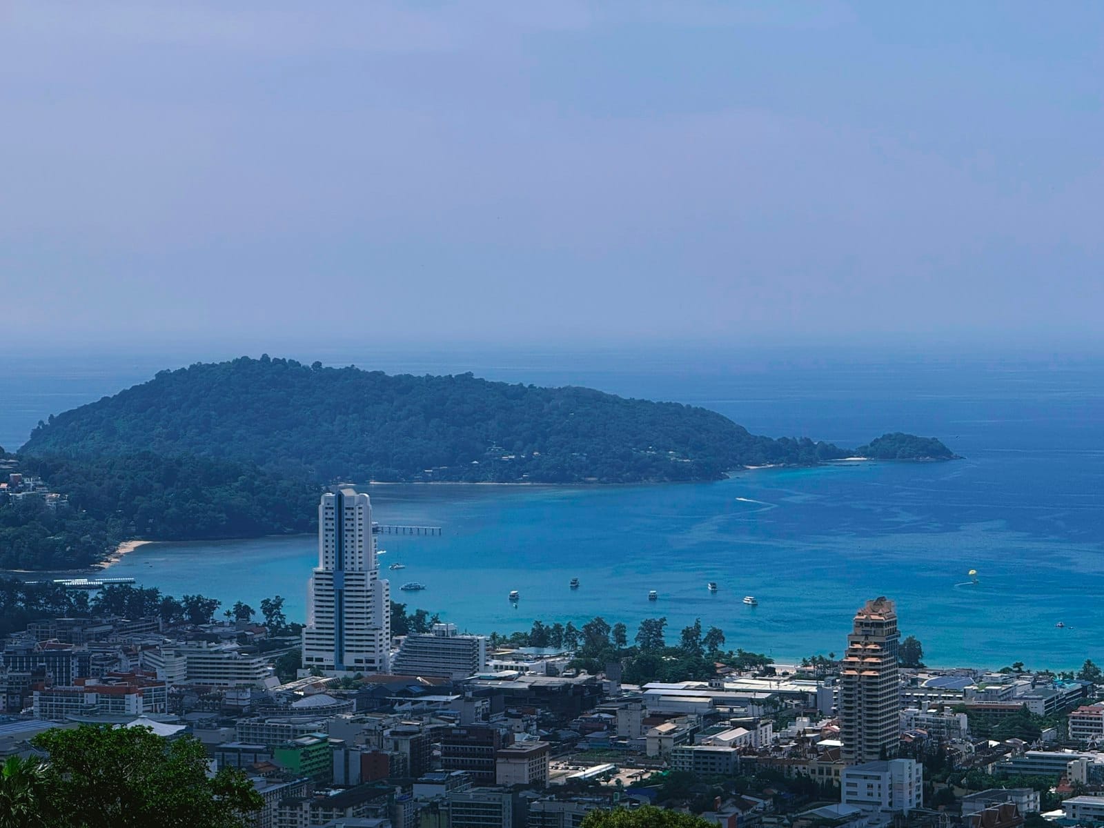 A panoramic view of Patong bay with turquoise sea, modern high-rise buildings, and a vibrant city stretching along the beach, under a clear blue sky.