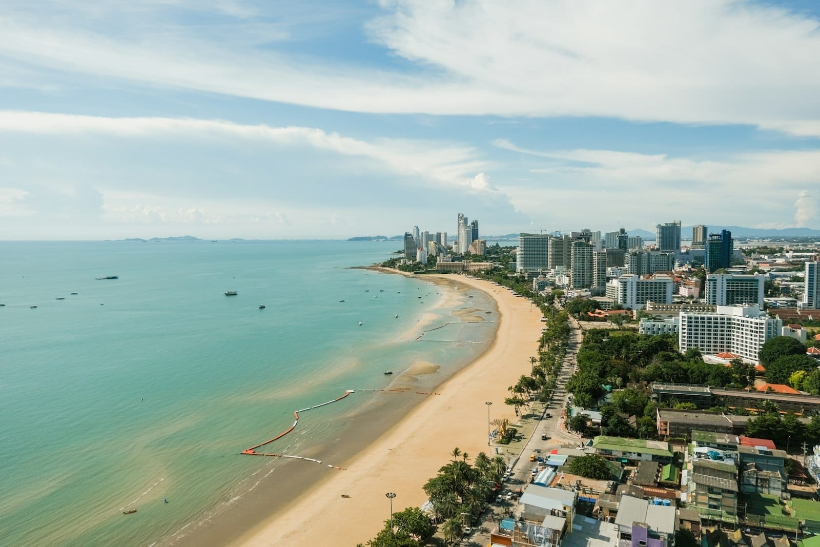 มุมมองทางอากาศของ Pattaya Beach แสดงให้เห็นทรายสีทอง น้ำสีเทอร์ควอยซ์ โรงแรมสูงริมชายหาด และถนนในเมืองที่คึกคักภายใต้ท้องฟ้าสว่างที่มีเมฆบางส่วน