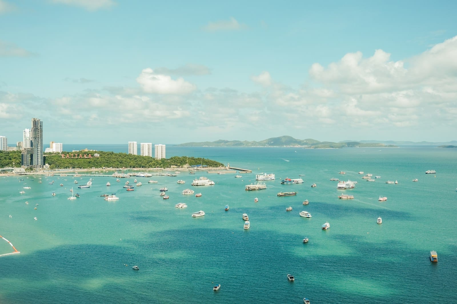 Vista aérea da Baía de Pattaya com barcos espalhados por águas turquesa, o horizonte da cidade e colinas verdes ao fundo sob um céu parcialmente nublado.
